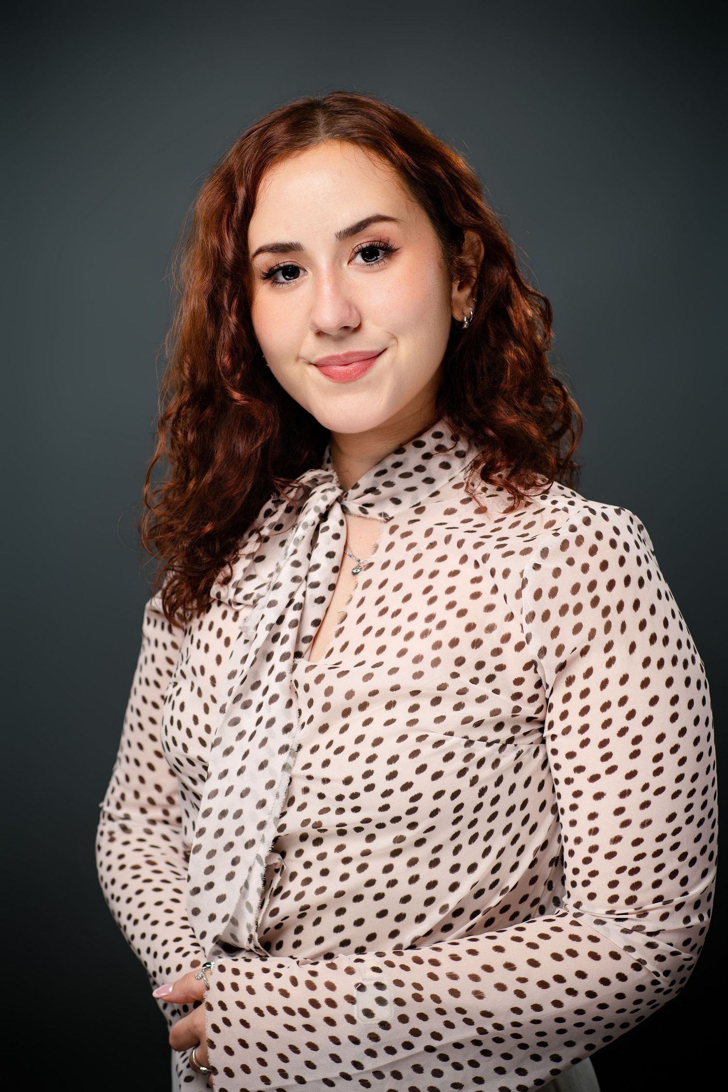 Young woman with curly red hair smiling, wearing a white polka dot blouse, posed against a dark gray background.