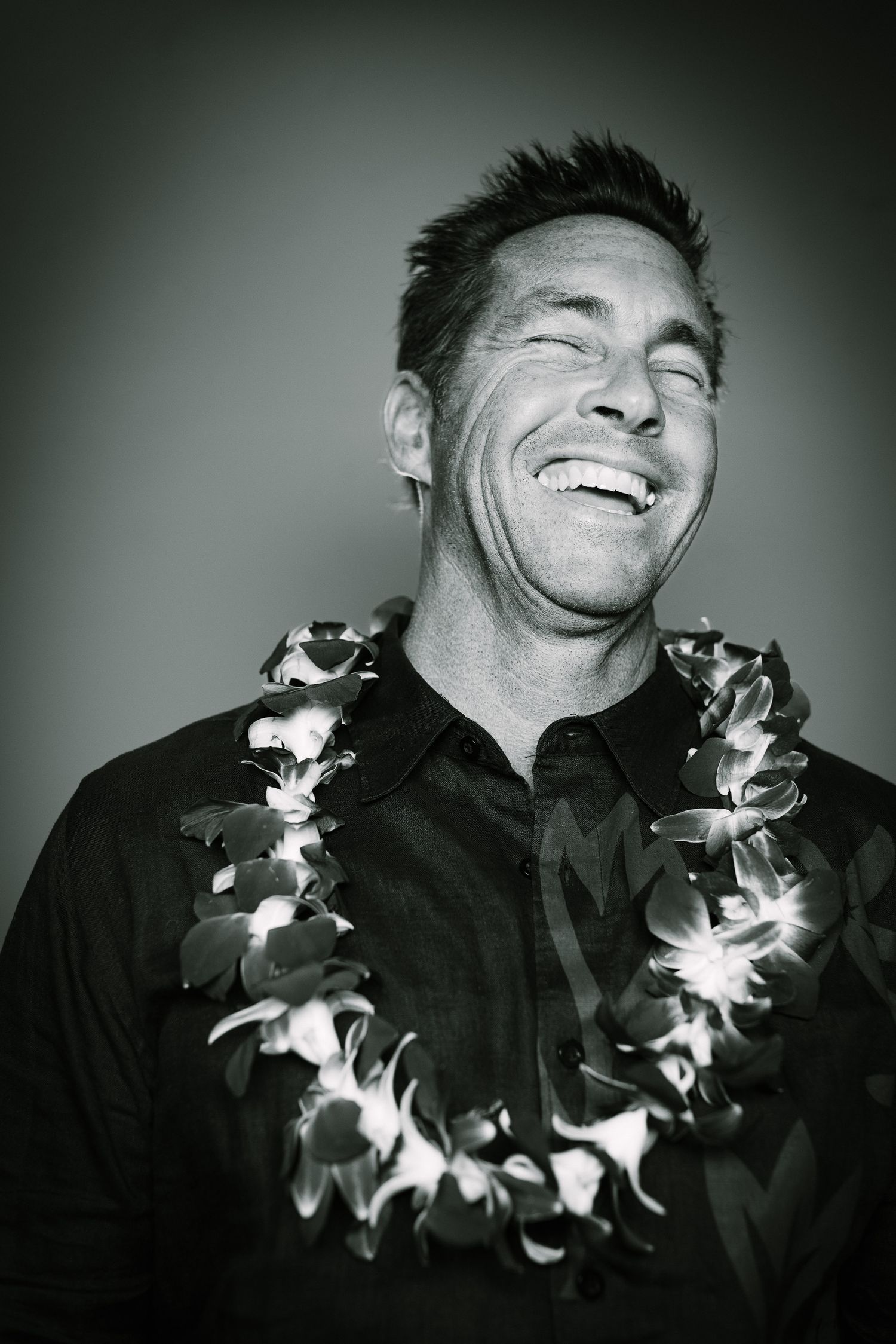Black and white candid portrait of a person with spiky hair wearing a lei and laughing joyfully.
