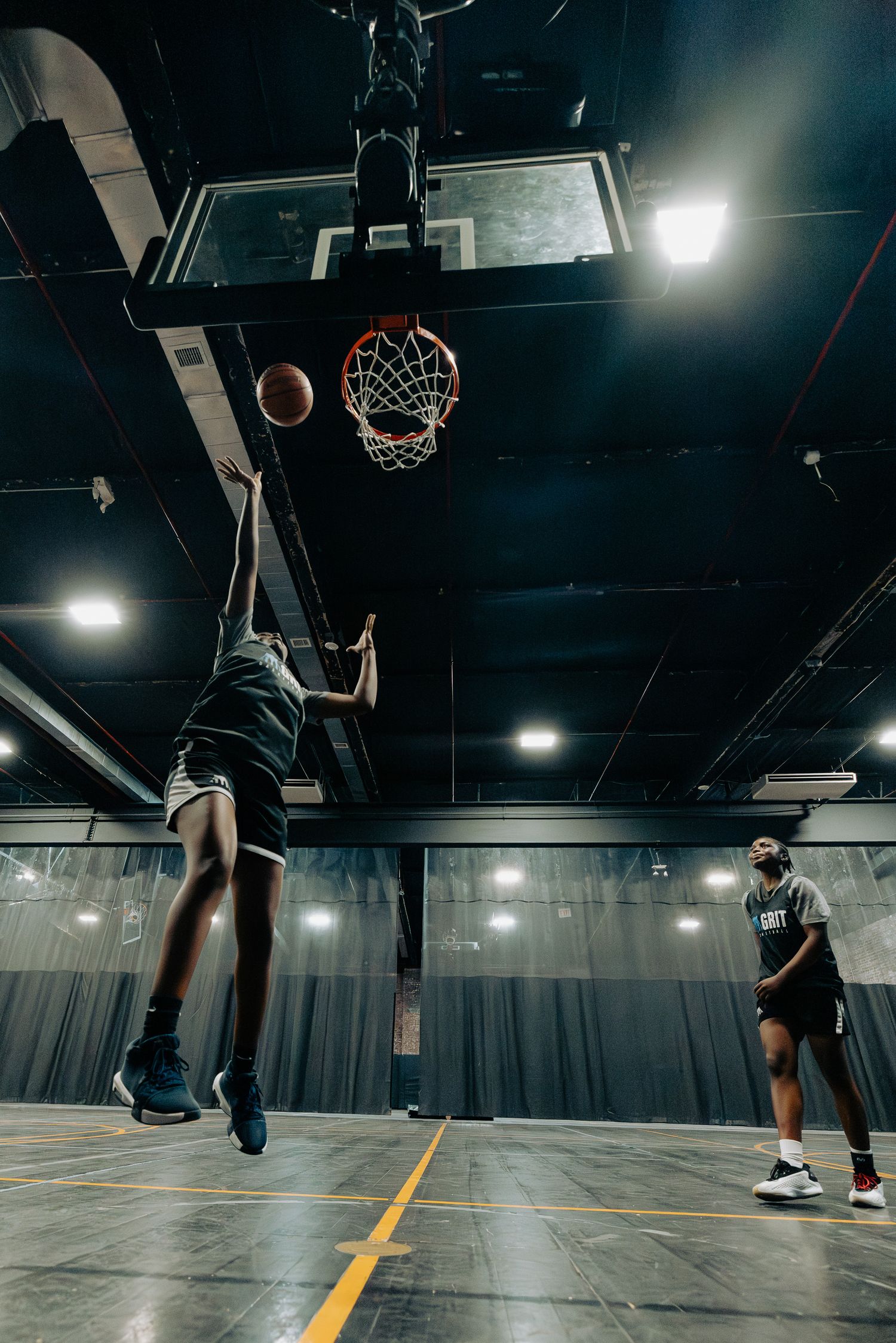 Basketball players practice on an indoor court with dramatic lighting as one player attempts a layup at the hoop.