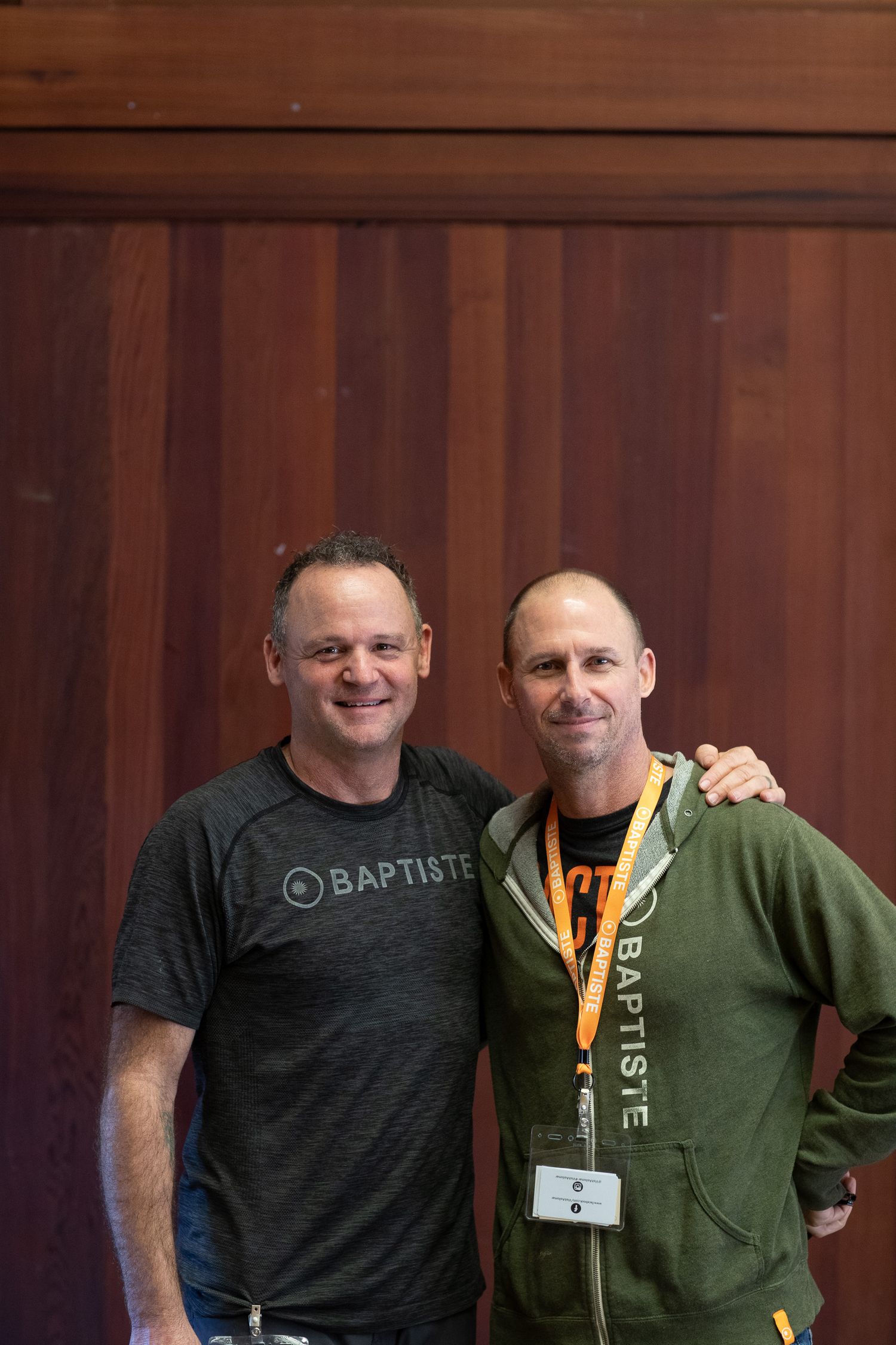 Two people smiling together against a wooden background wearing casual athletic attire.