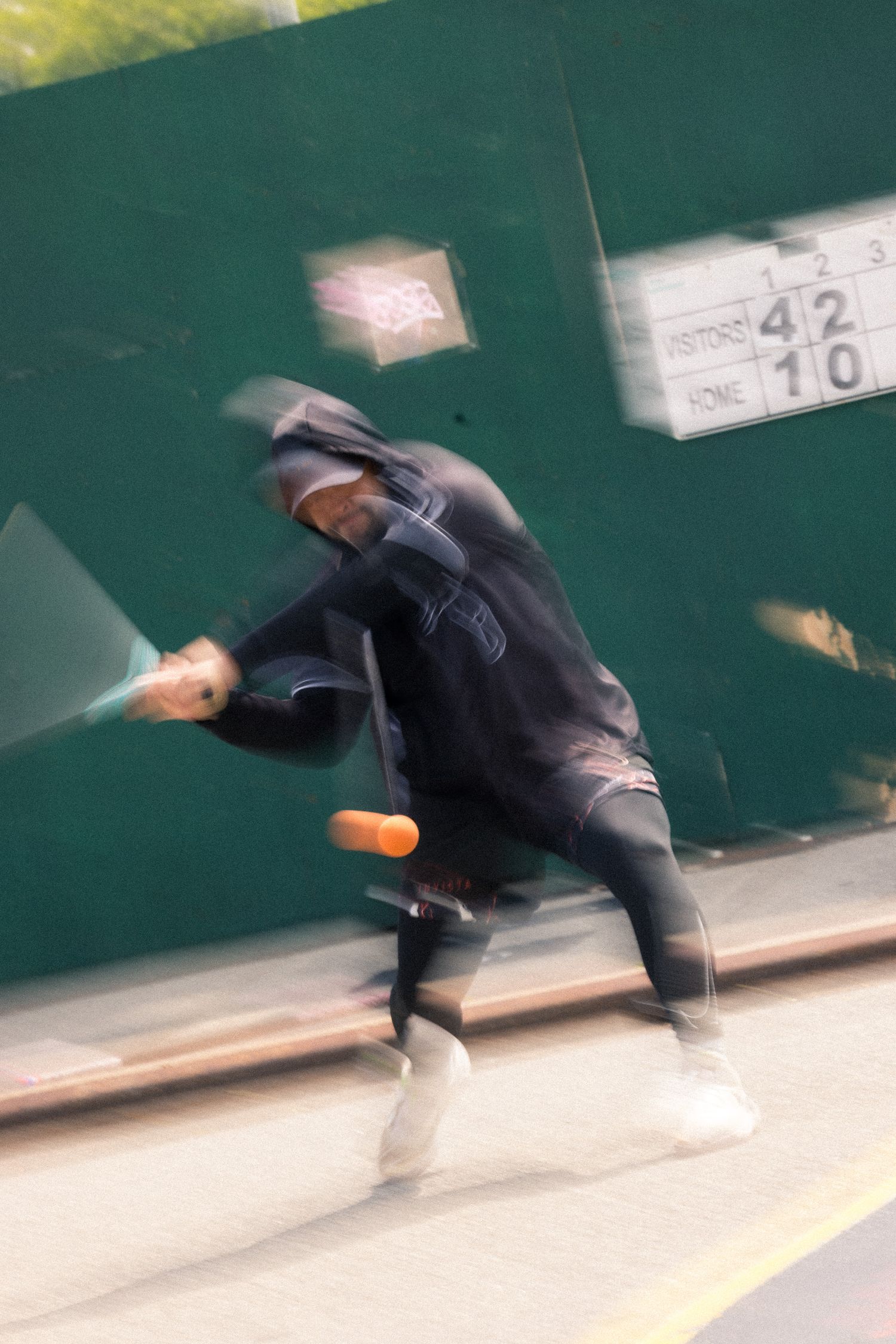Motion blur captures person in black clothing performing dynamic dance moves against a green wall.