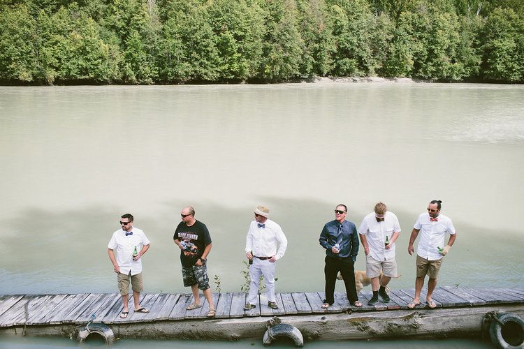 Wedding party stands on long wooden dock overlooking water.
