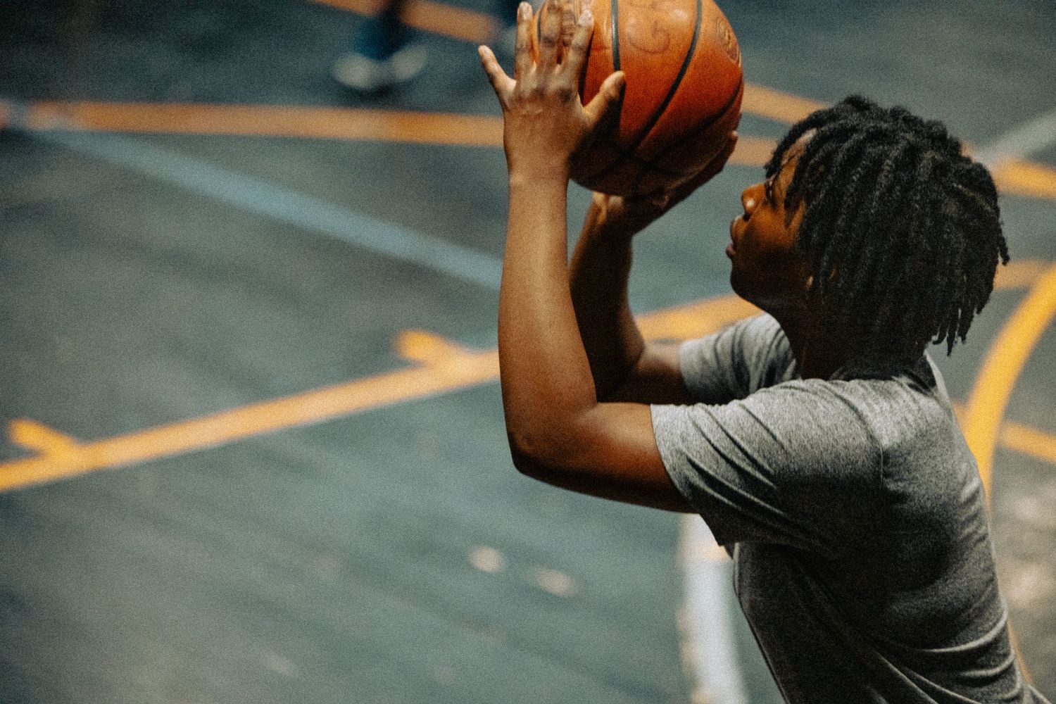 Basketball player in grey t-shirt practicing shooting form with ball raised over head in indoor gym.