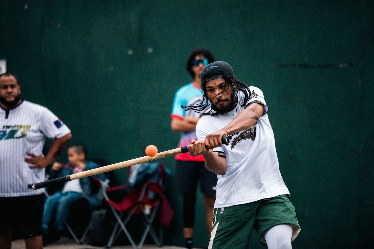 A player swings a bat during a street baseball game against a green wall background.