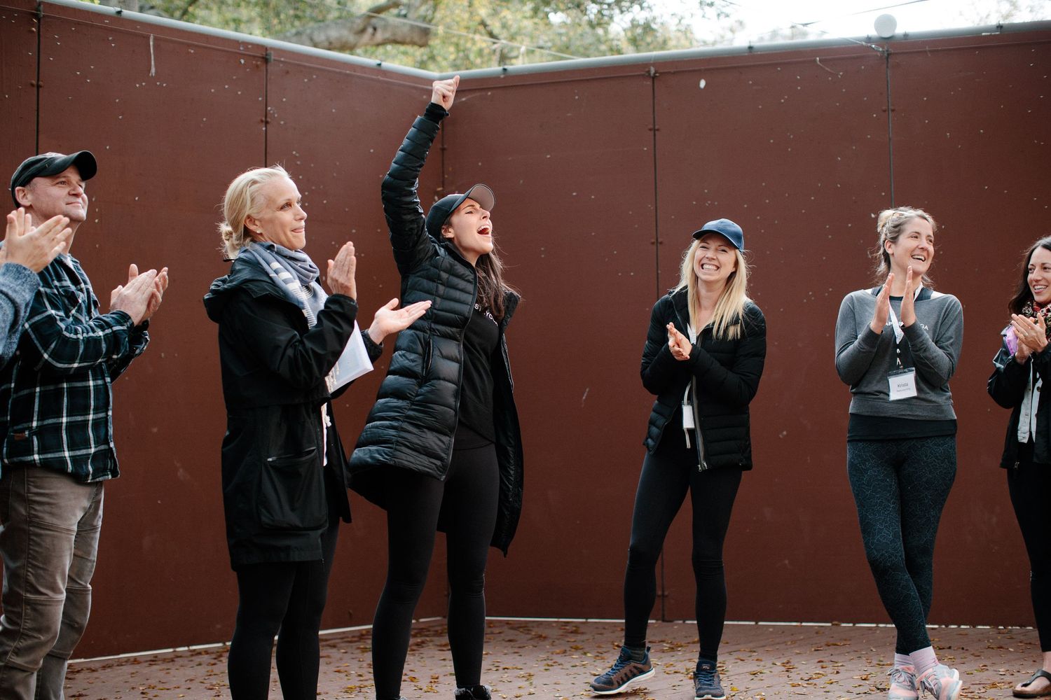 A group of people in winter clothing raise their hands and laugh together against a rusty brown wall.
