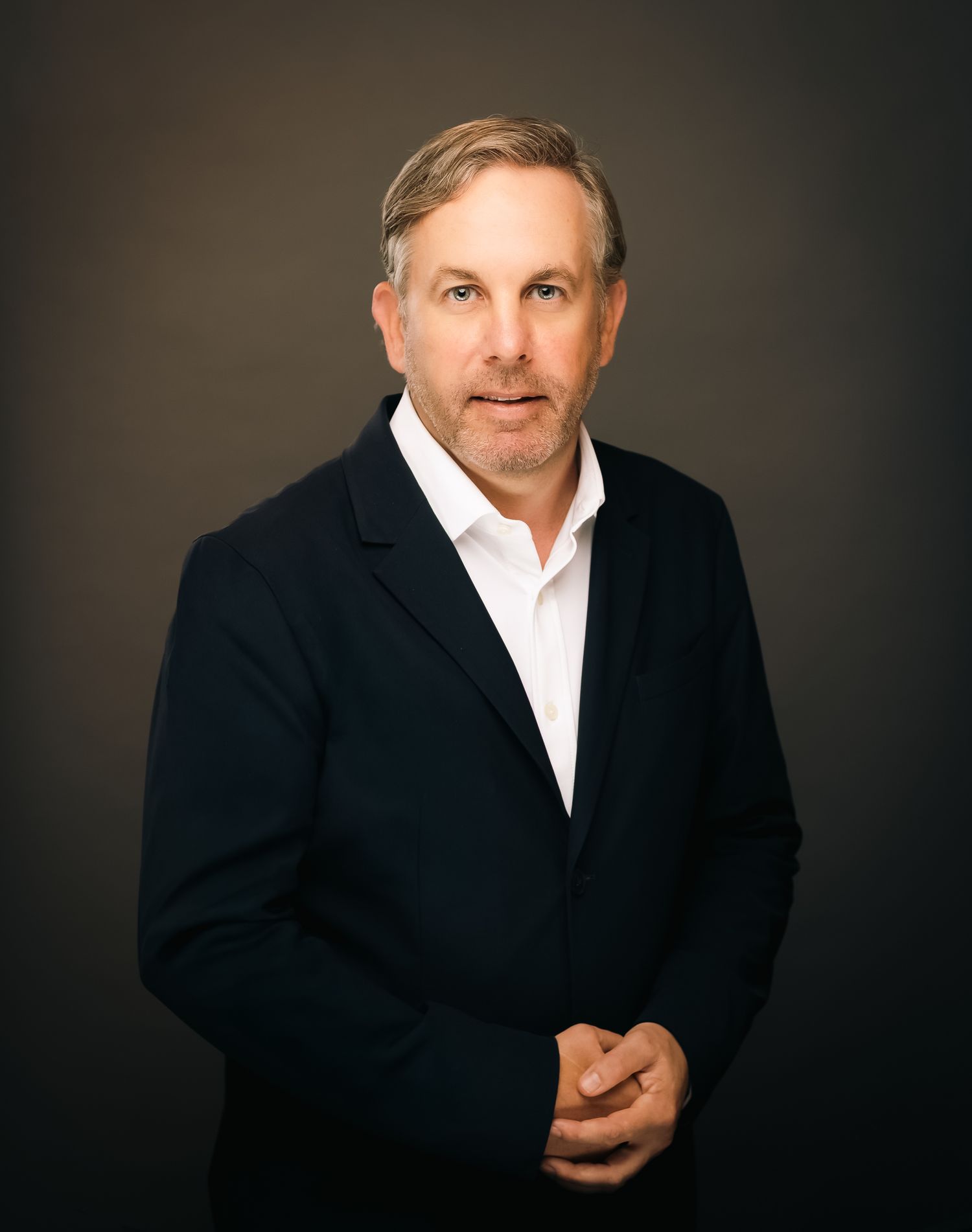 Professional headshot of a middle-aged man in a navy blazer and white shirt, posing confidently against a dark background.