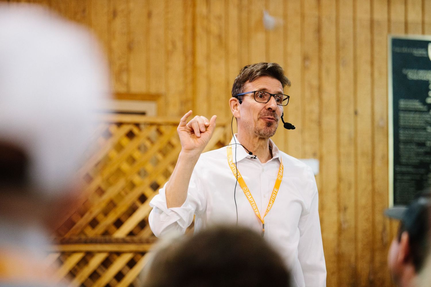 A speaker wearing glasses and a white shirt gestures enthusiastically during a presentation in a wood-paneled room.