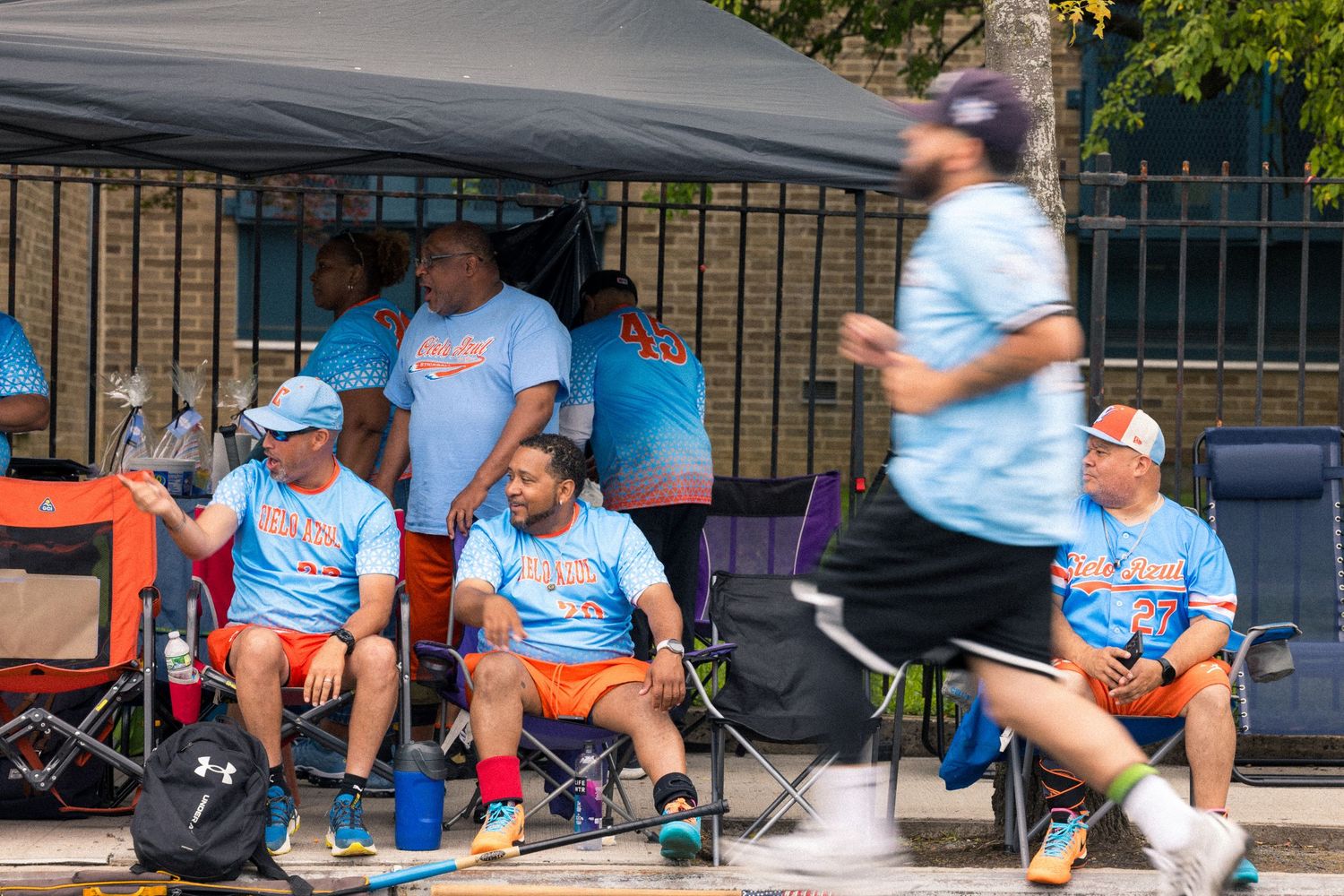 Baseball fans in matching light blue shirts sit under a canopy tent at an outdoor sporting event.