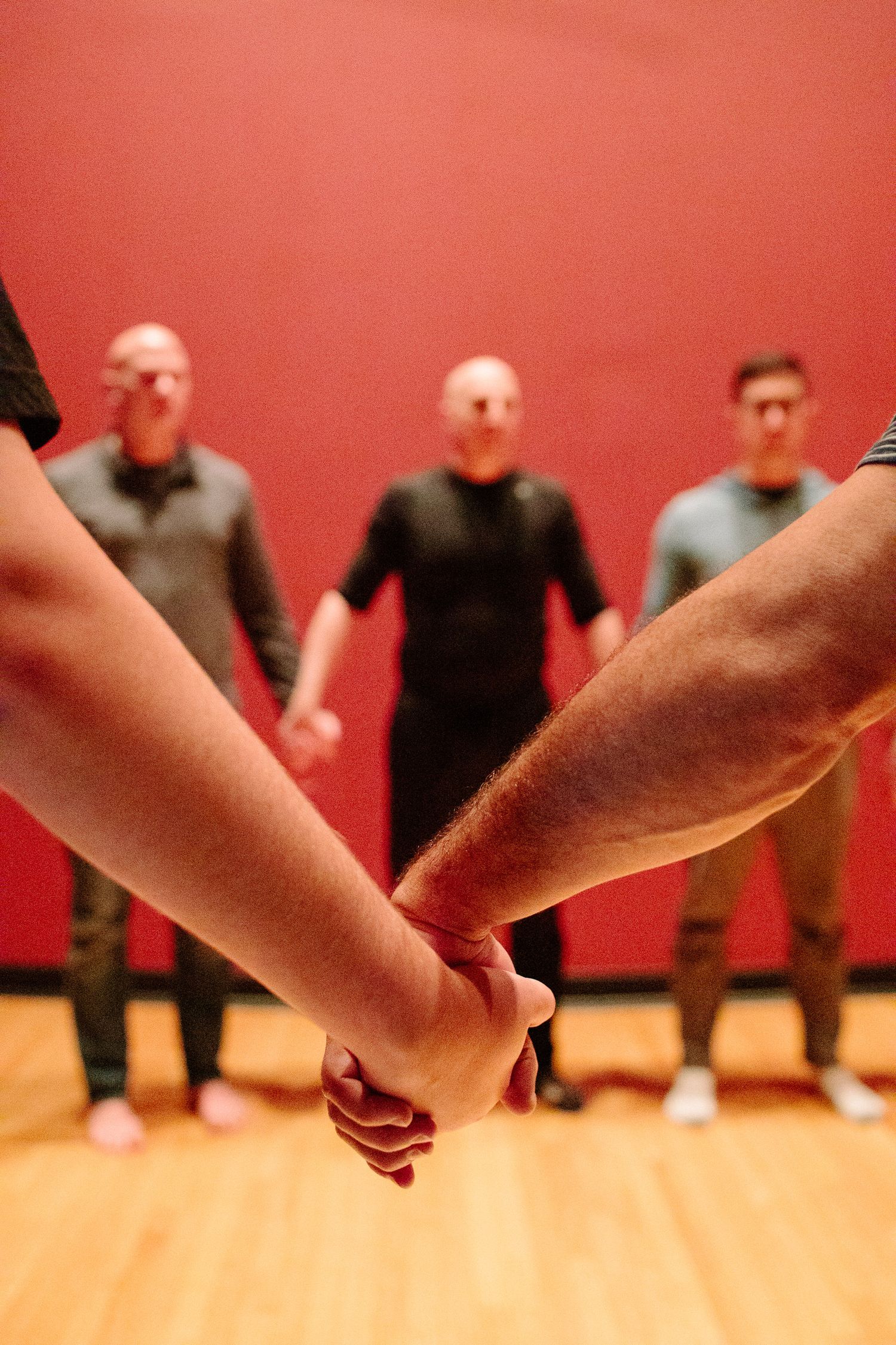 People hold hands in a circle on a wooden floor against a red wall during a group activity.