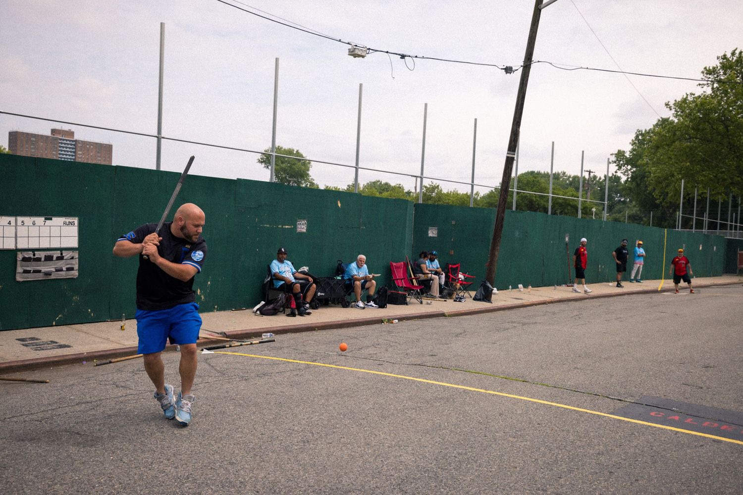 Players practice stickball on an urban street court with a green wall and spectators watching from the sidelines.