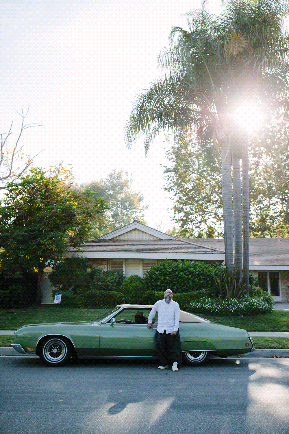 A green vintage Buick Riviera parked on a suburban street beside a house with palm trees in the late afternoon sun.