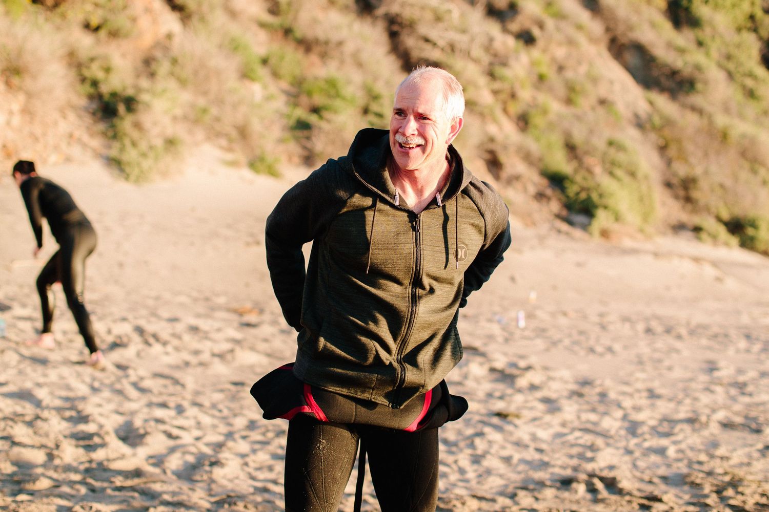 People walk and exercise on a sandy beach with hills in the background.