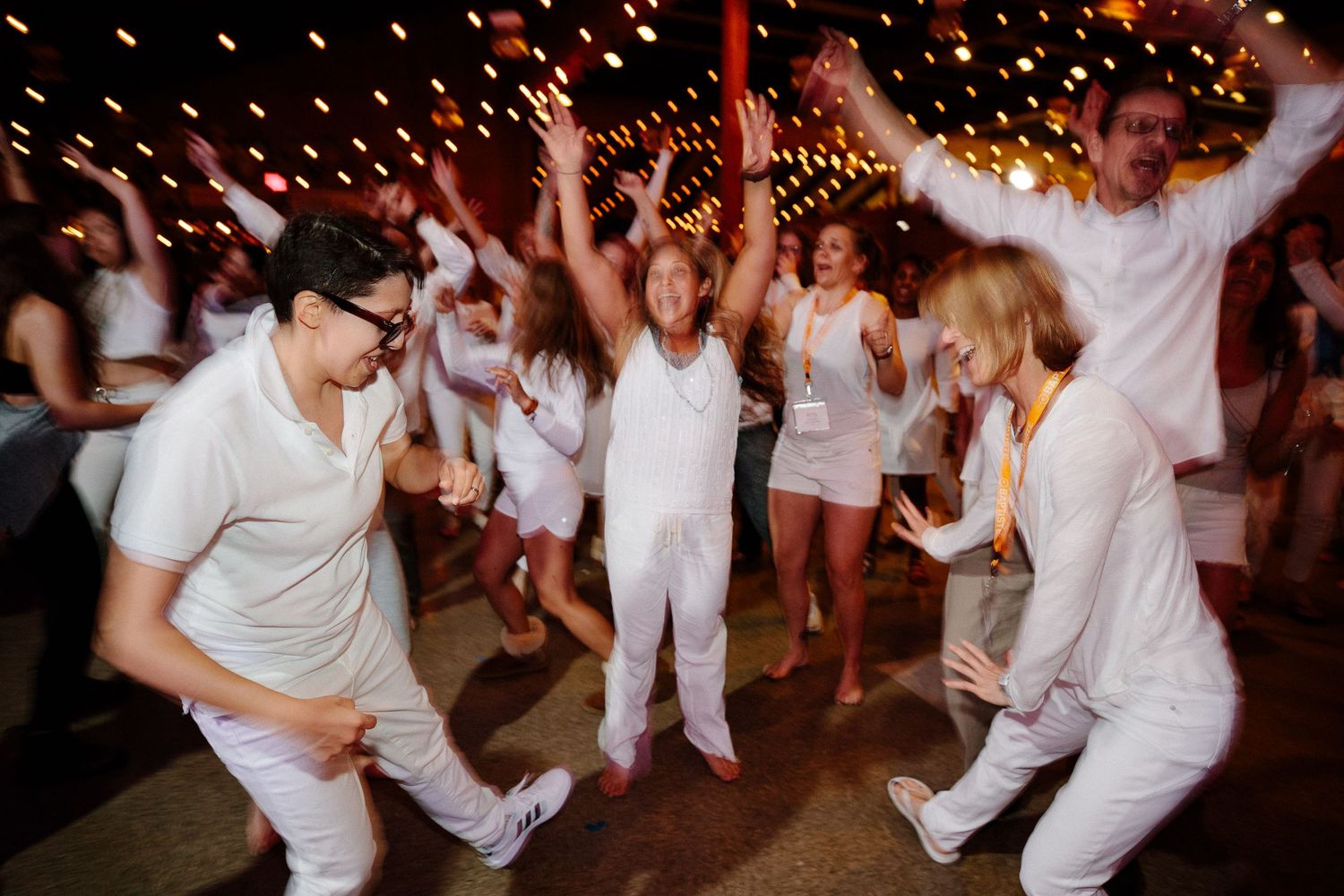 Dancers in white clothing moving energetically under sparkly lights at a nighttime event.