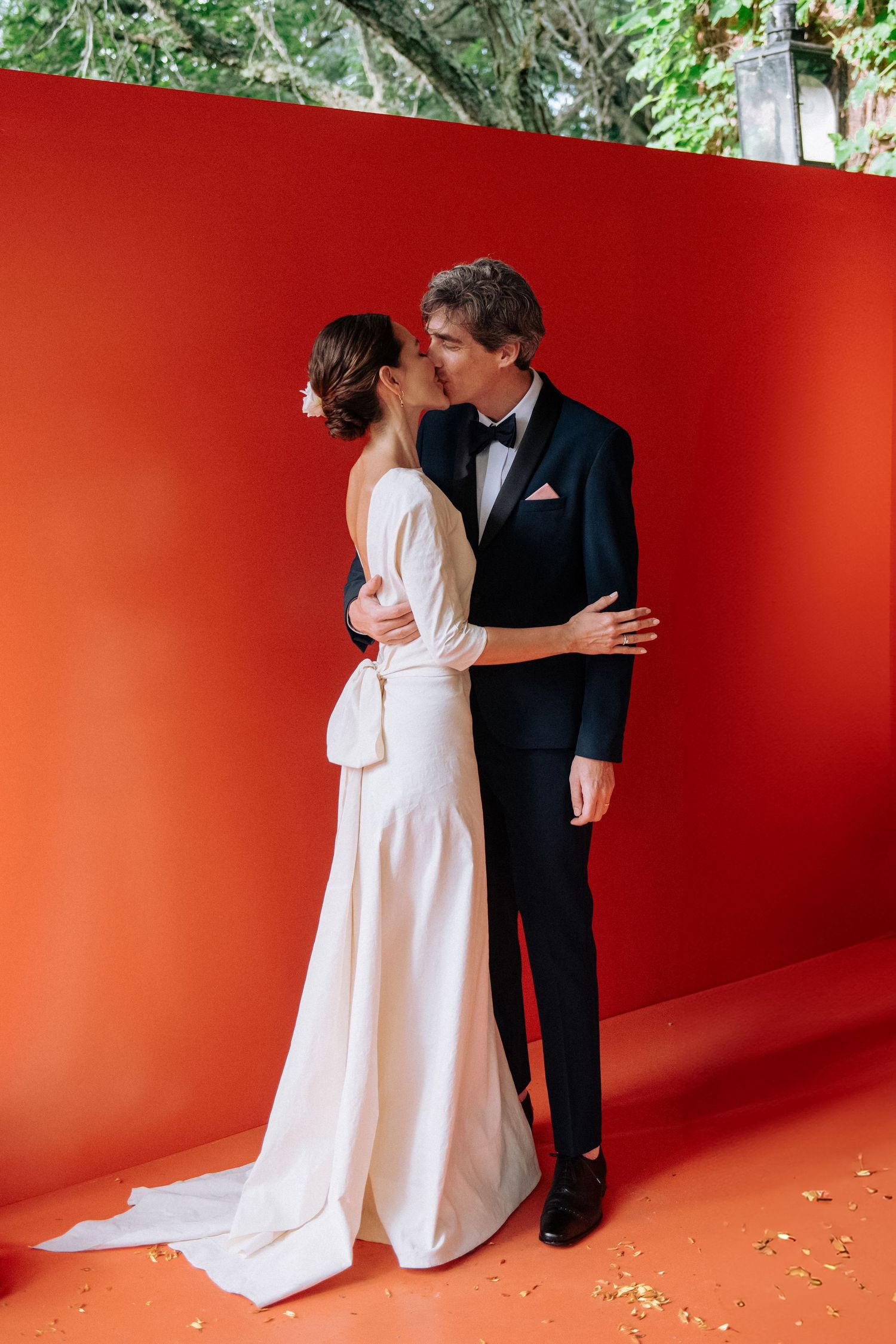 A romantic couple in formal wedding attire embracing against a red wall with greenery visible in the background.