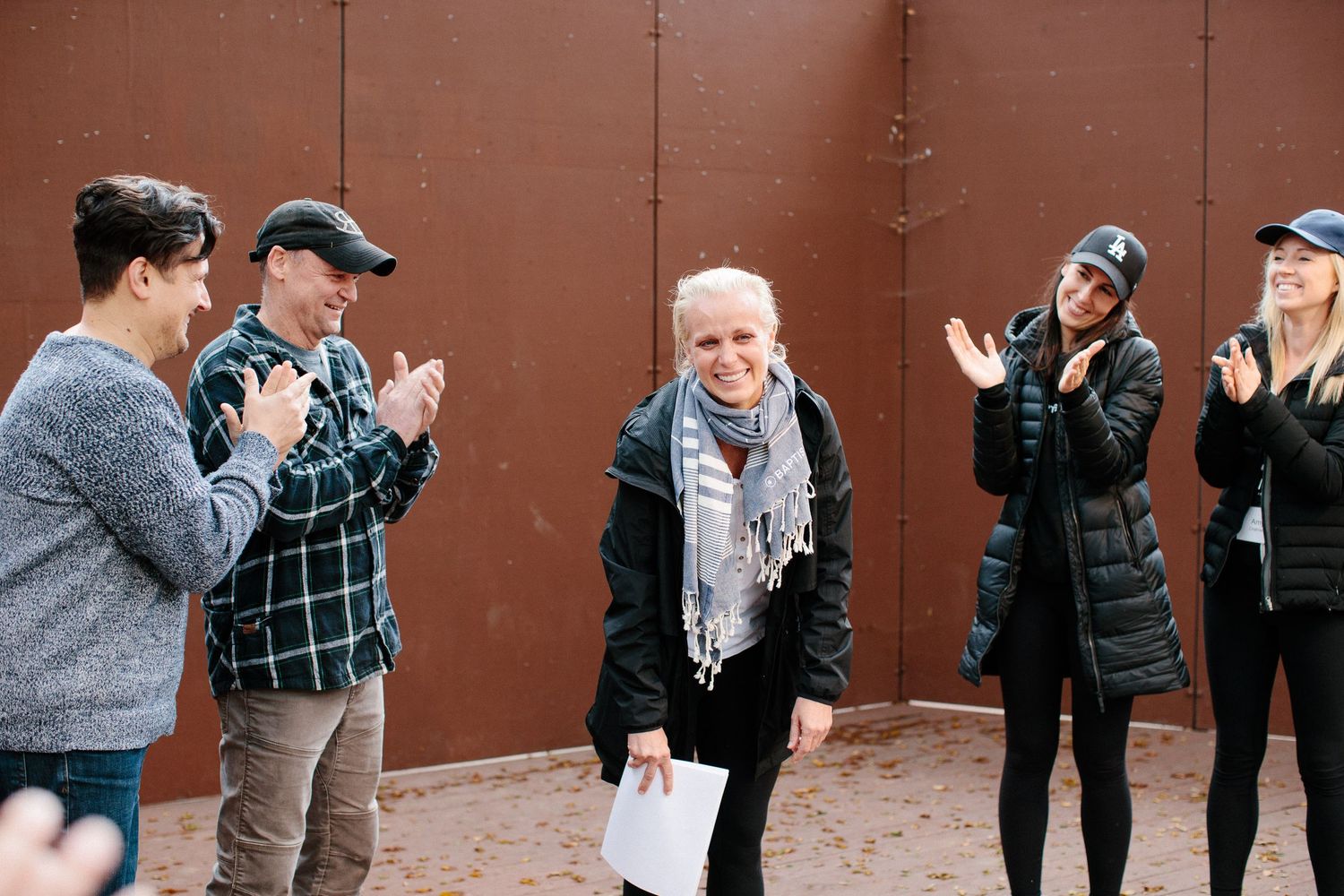 People in casual winter attire conversing and standing by a brown wall outdoors.