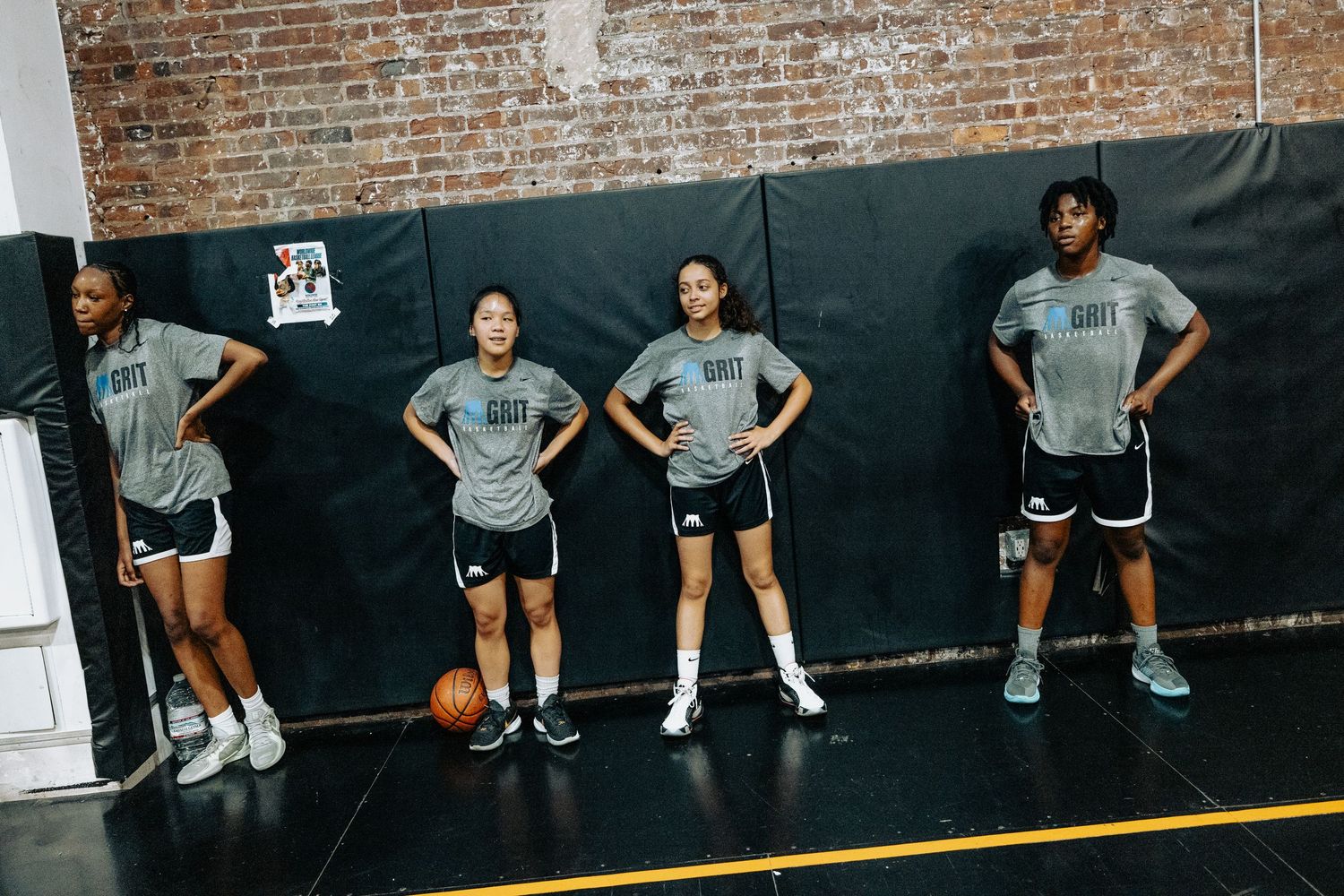 Basketball team lines up against wall during practice drill in brick-walled gym.