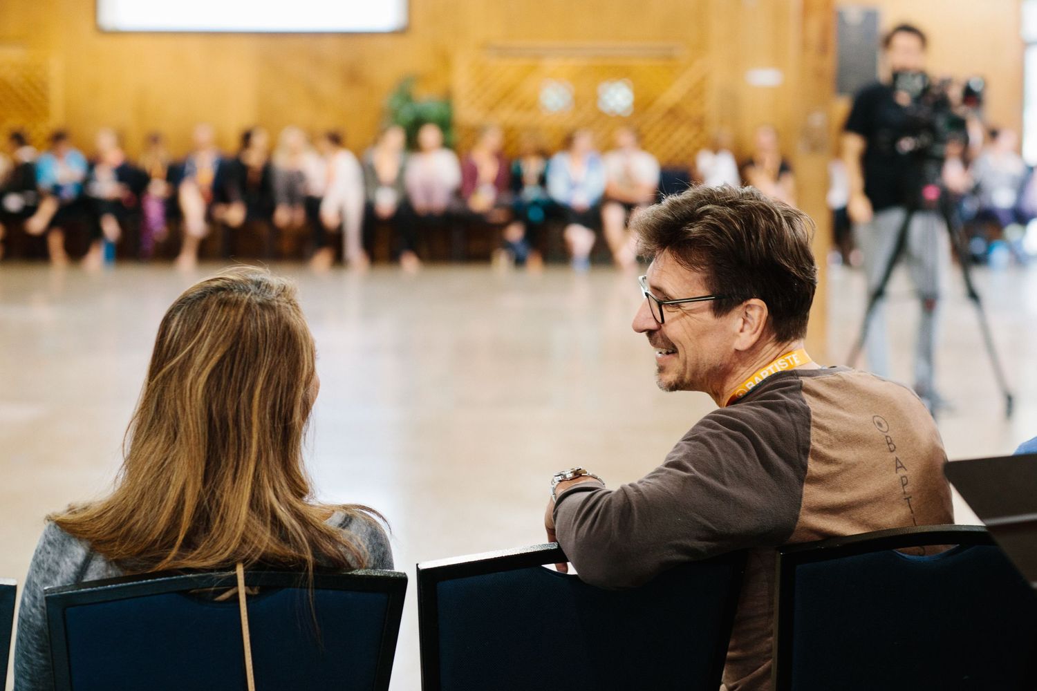 Two people engage in conversation while seated in chairs at indoor venue with wooden wall backdrop.