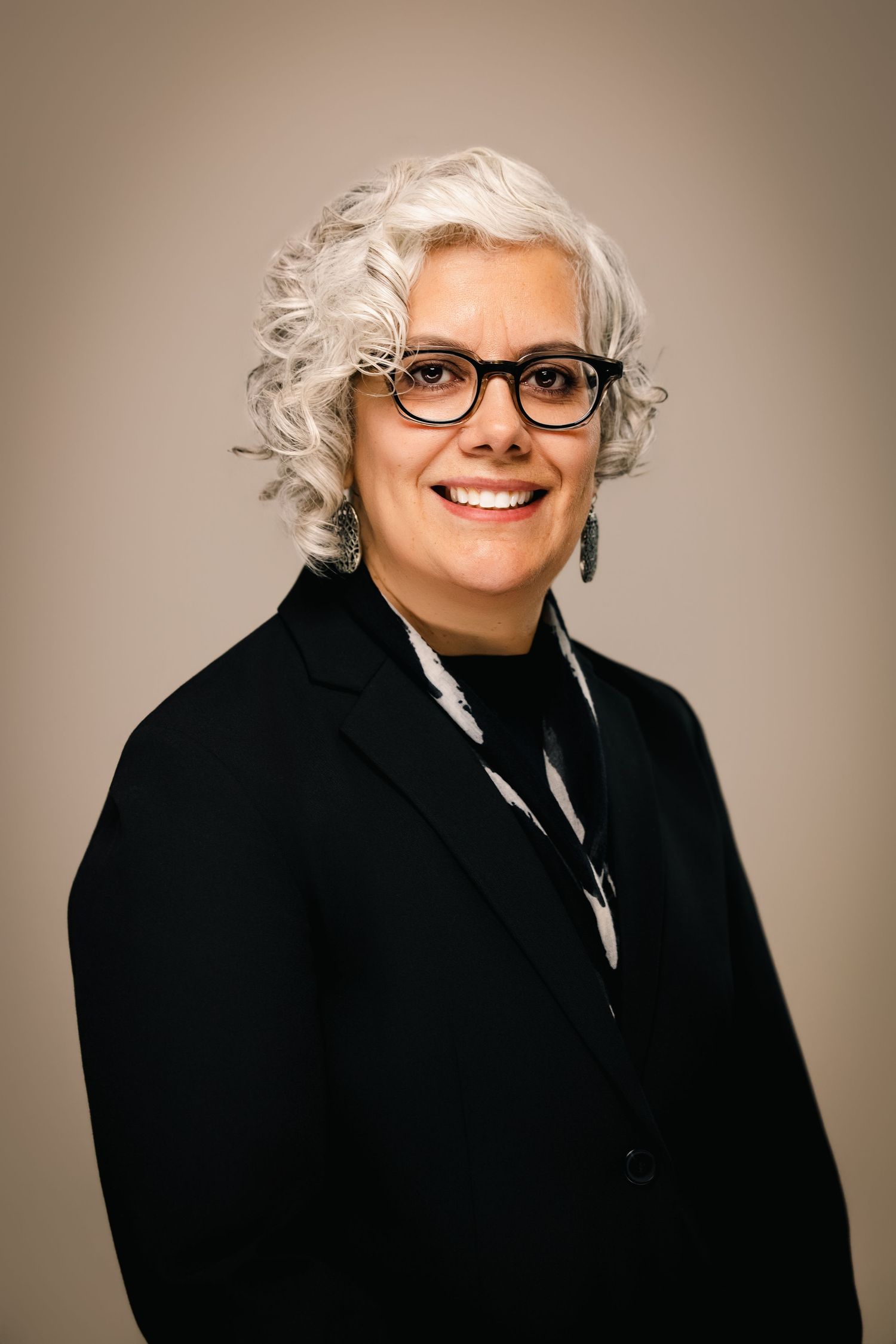 Professional woman with white curly hair and black glasses wearing a black blazer, smiling confidently for a headshot.