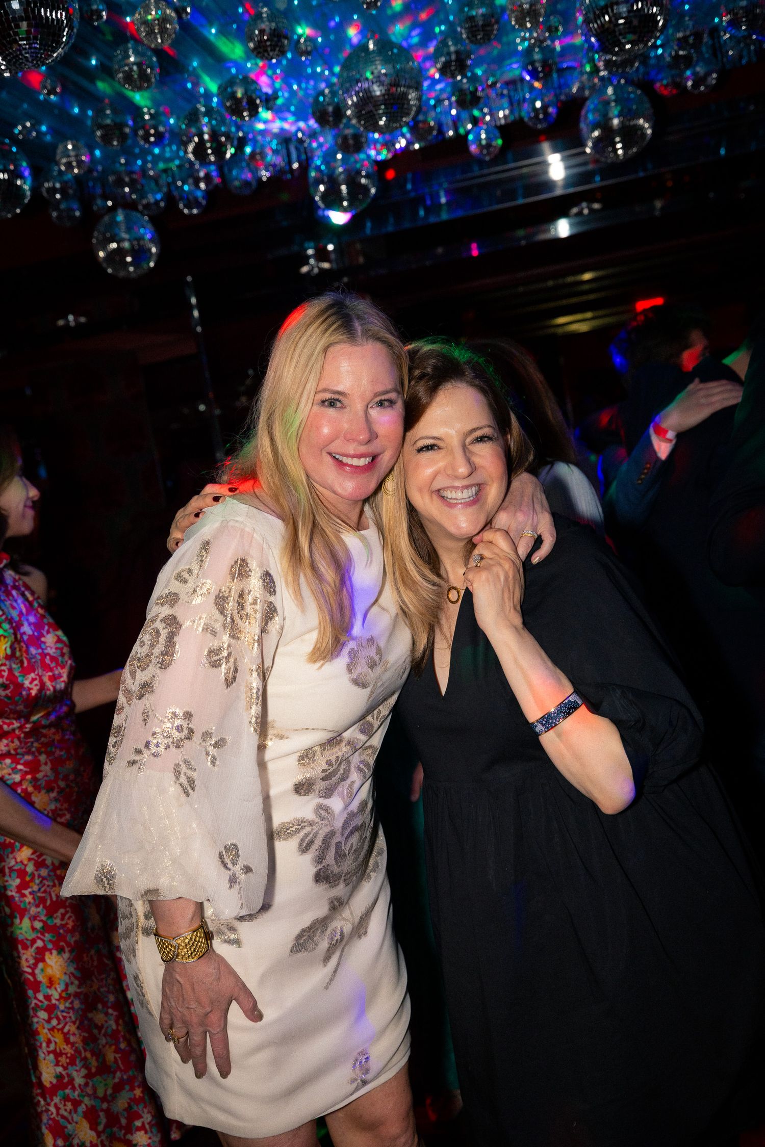 Two friends embrace and smile under colorful disco lights at a nightclub party.