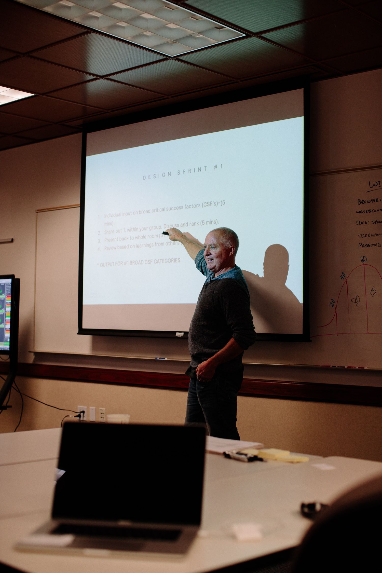 Presenter giving a lecture in front of projection screen in conference room setting.