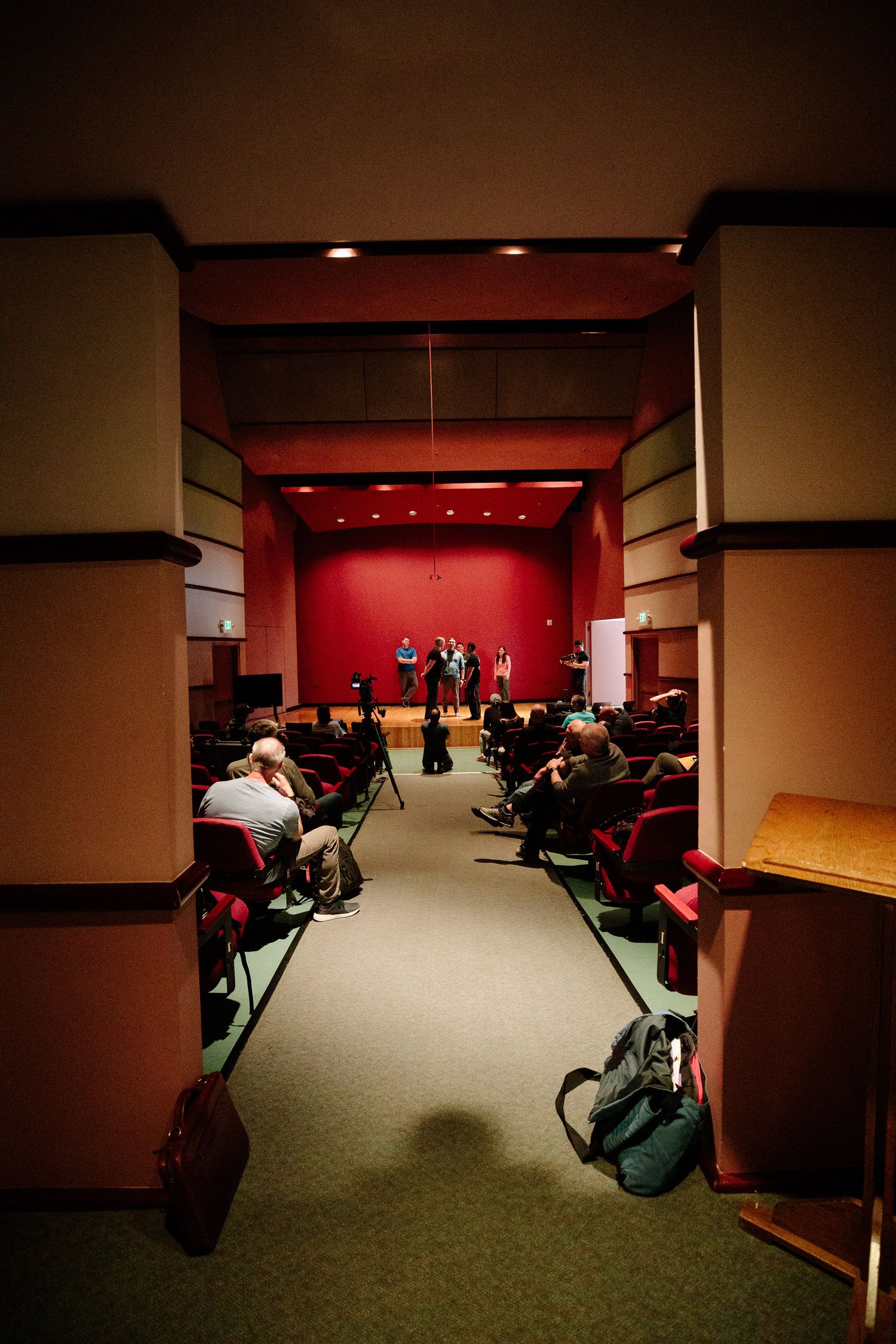 View down a theater aisle with red walls and audience members seated in chairs.