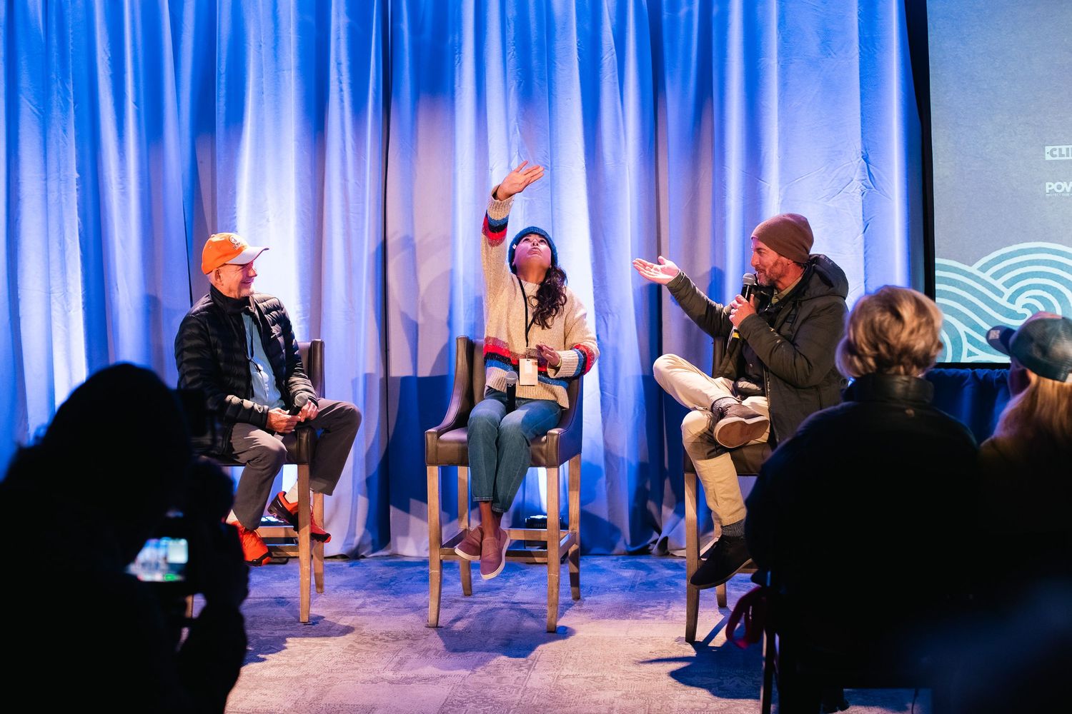 Panel discussion on stage with blue curtain backdrop and audience silhouettes.