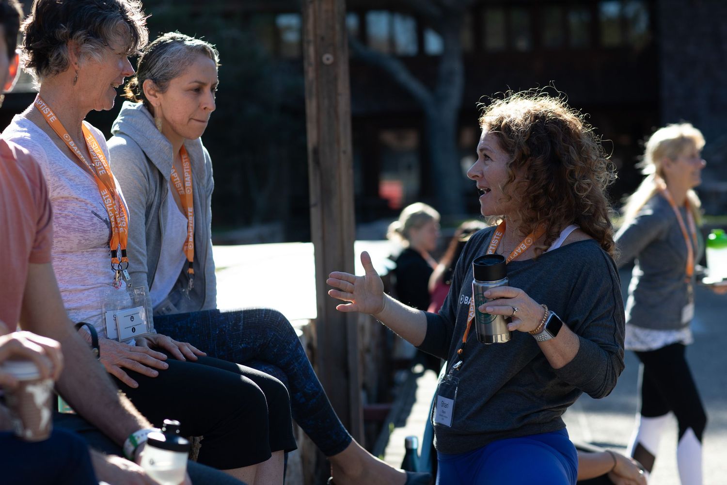 Three people engaged in an outdoor discussion on a sunny day with other participants visible in background.