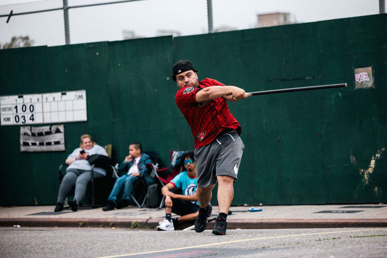Player in red shirt practices stickball swing form against green wall while spectators watch in background.