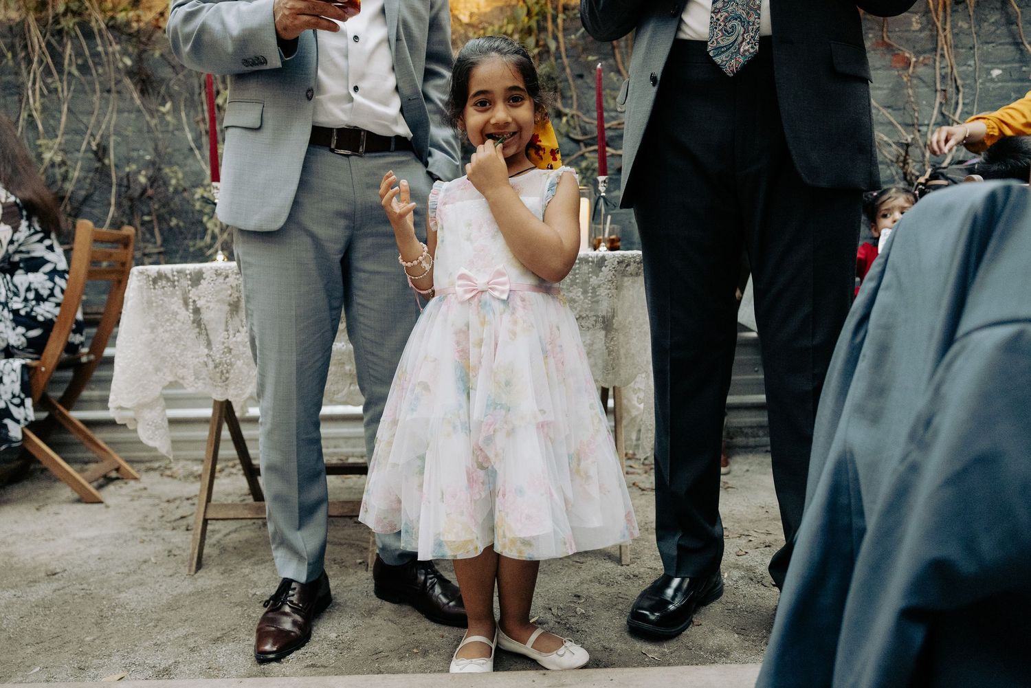 A young girl in a pink party dress stands between two adults at an outdoor gathering.