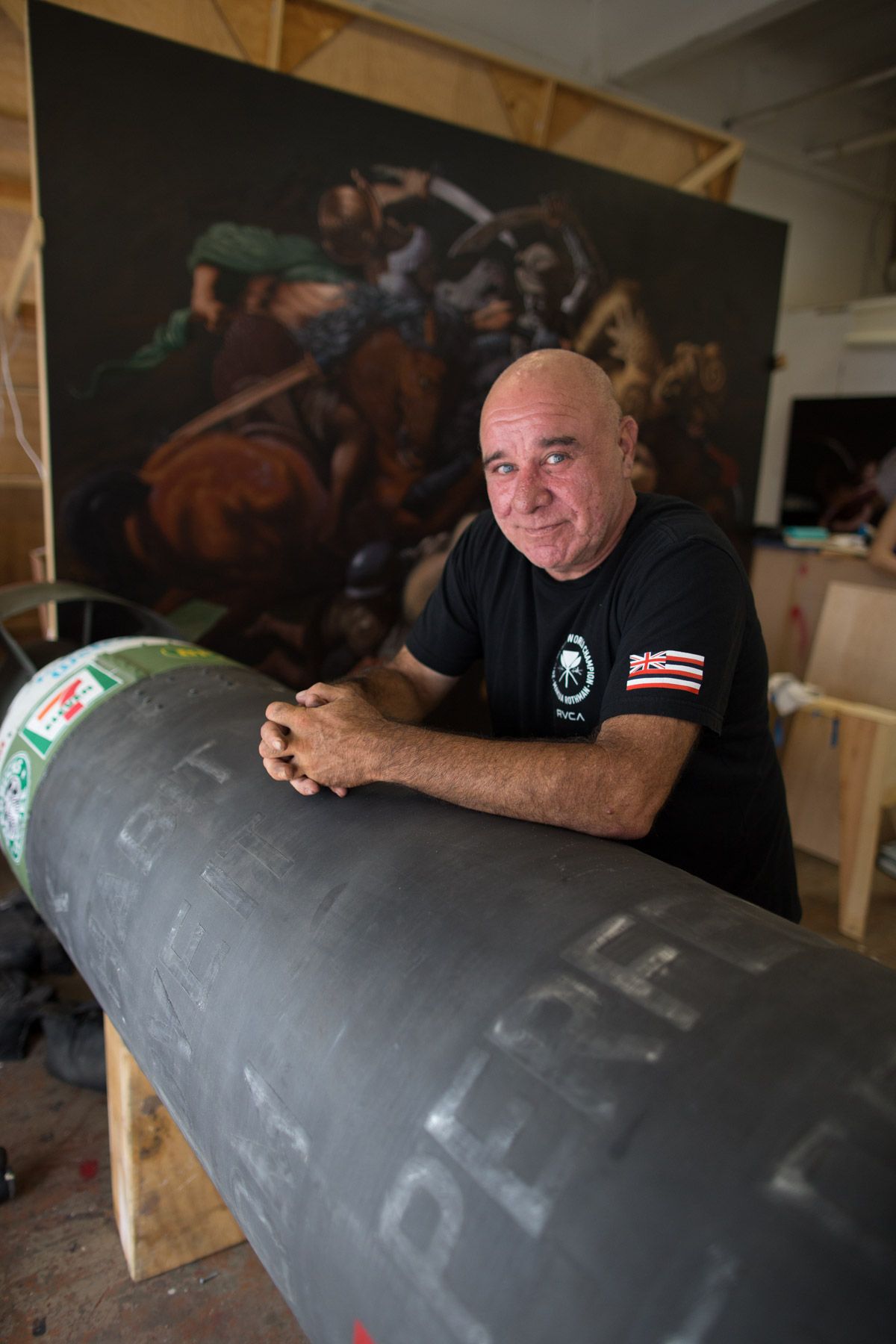 Person in black shirt leans on large cylindrical object with dramatic painting in background.