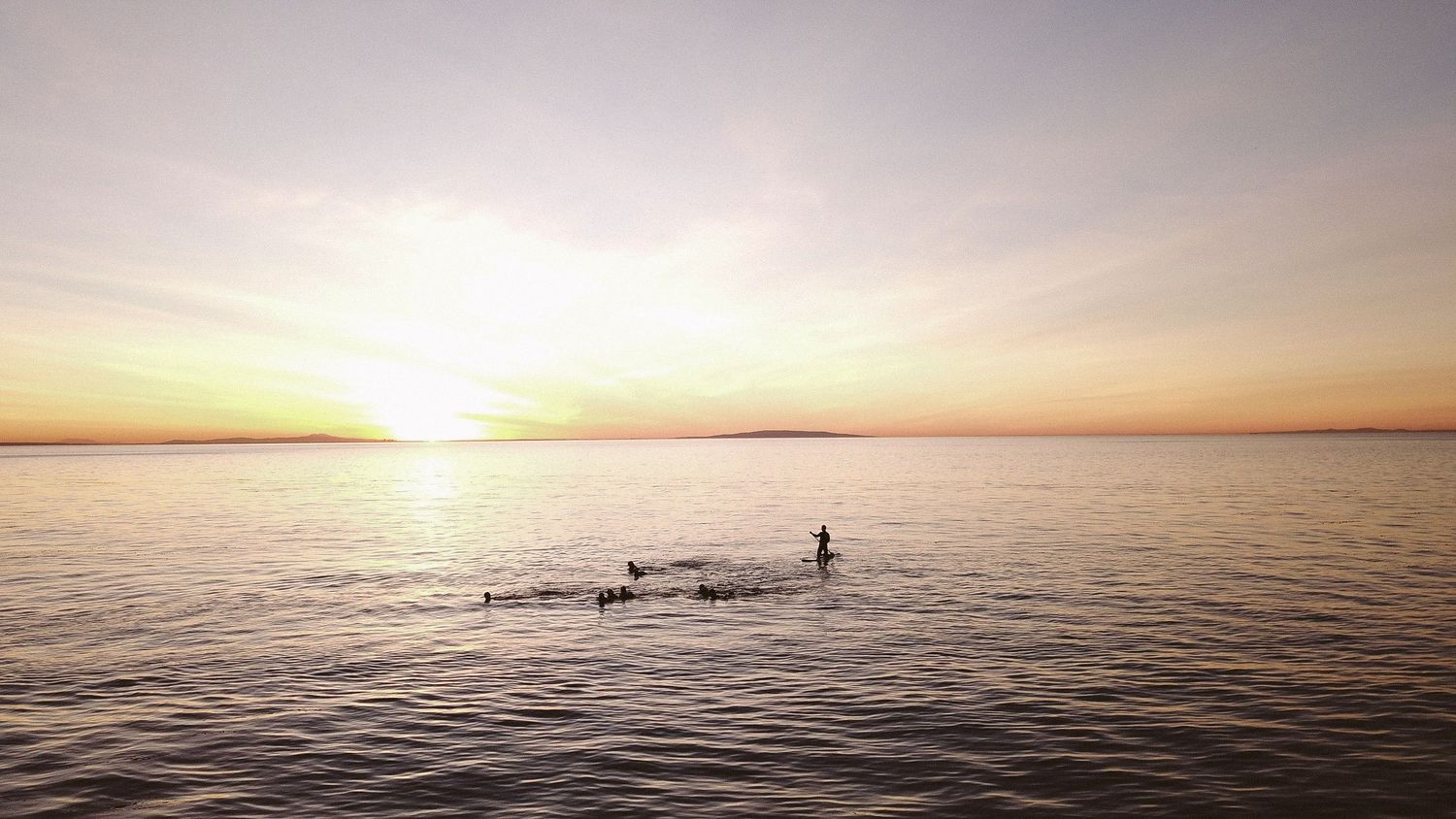 Panoramic view of a calm ocean at sunset with distant swimmers.
