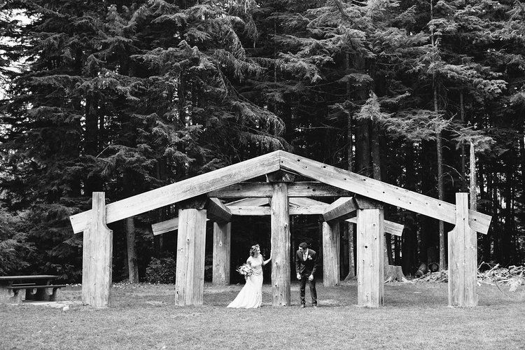 A black and white photo of a rustic outdoor wedding ceremony taking place under a wooden pavilion in a forest setting.