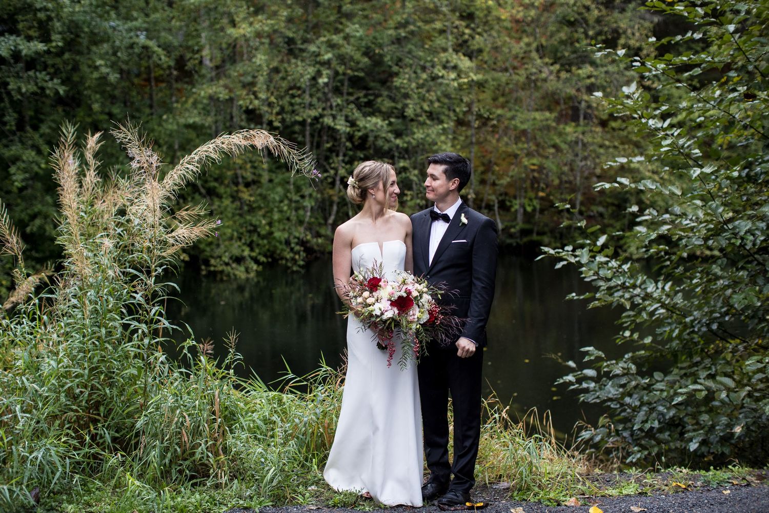 A couple in wedding attire stands together on a path surrounded by lush green foliage and natural scenery.