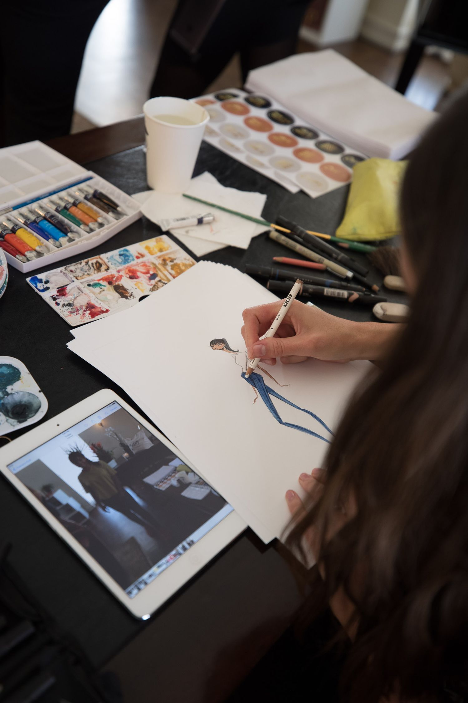 Artist sketching at desk with watercolor supplies and reference photos.