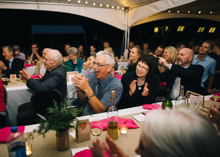 Wedding reception guests applauding during evening celebration under tent lighting.