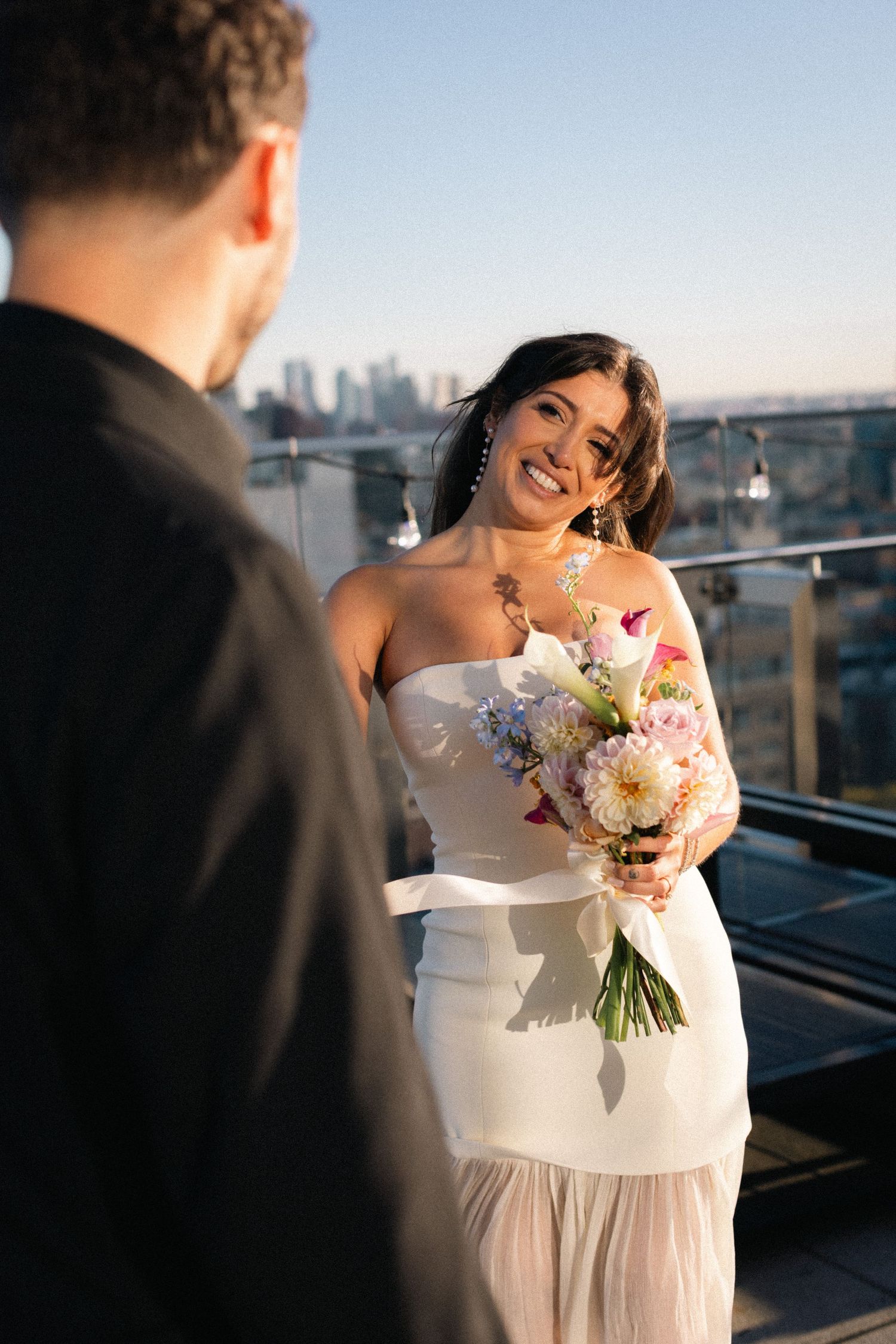 Smiling bride in white strapless gown holds colorful bouquet on rooftop with city skyline at golden hour.