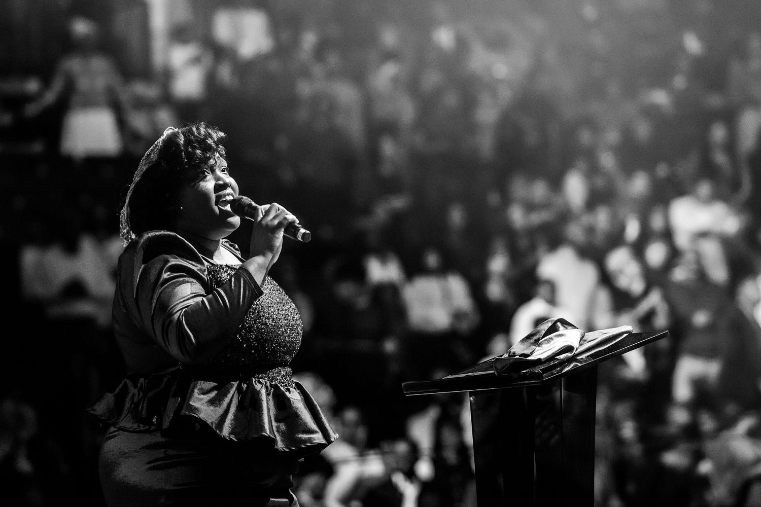A dramatic black and white concert performance photo showing a silhouetted performer on stage with a crowd in the background.