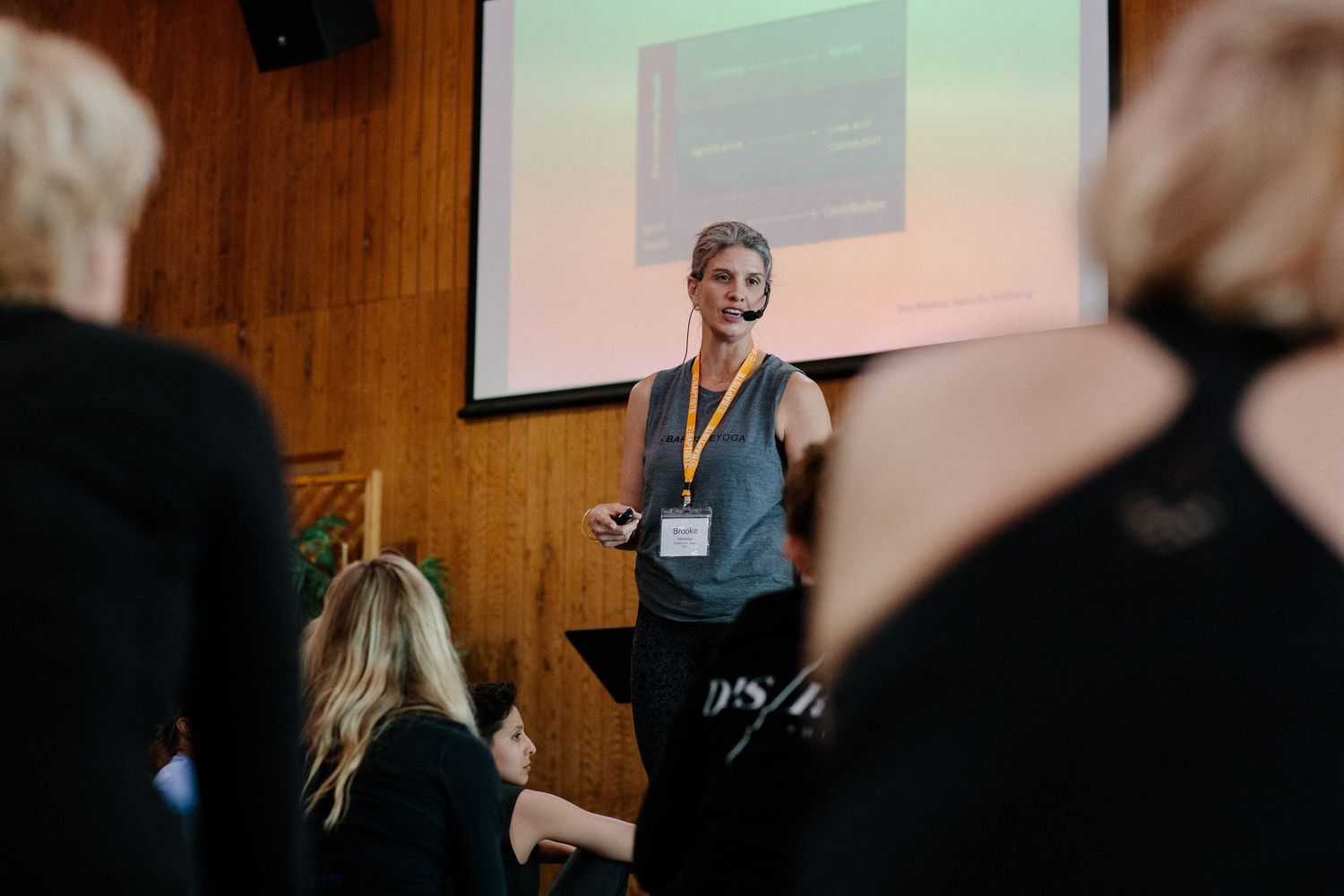 A presenter stands in front of a projection screen during a conference presentation, wearing business attire with a lanyard.