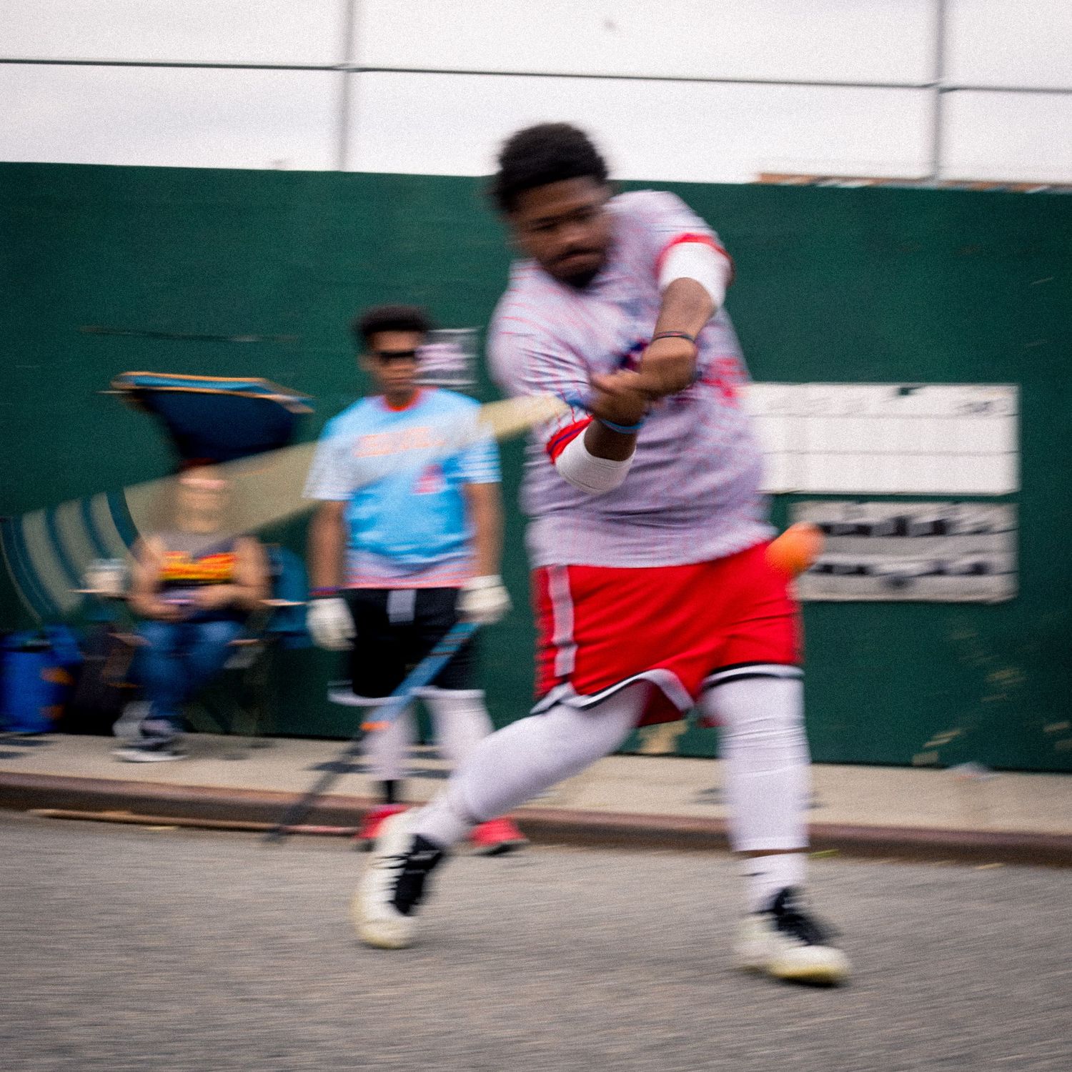 Baseball player in red shorts swings a bat during a street game with motion blur effect.