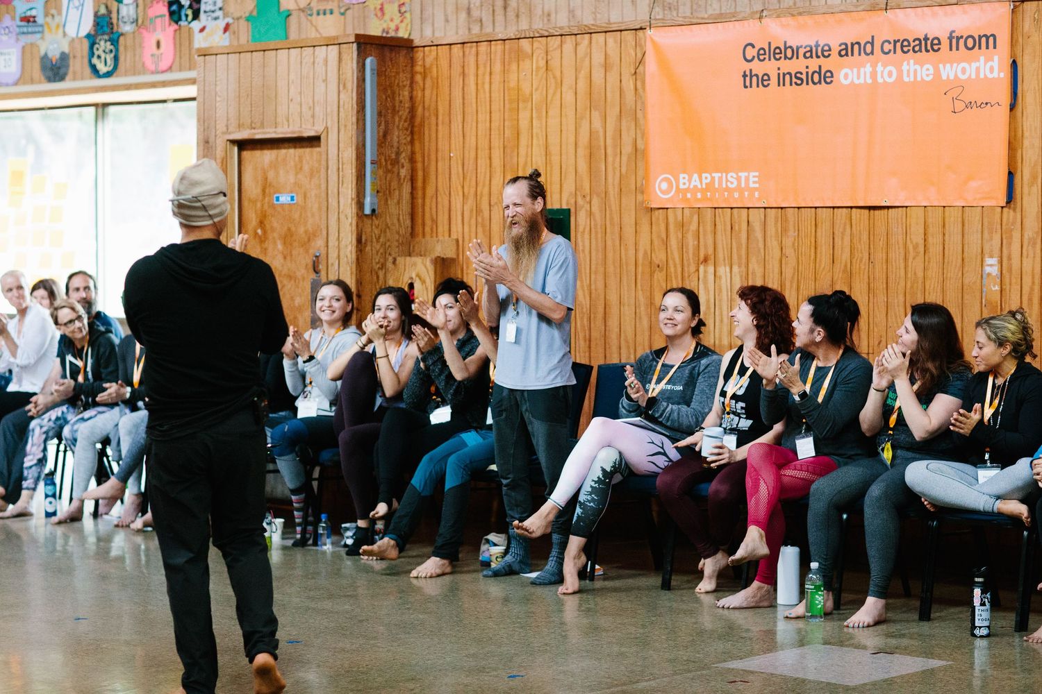 People seated along wooden walls with bright orange banners during an indoor event.