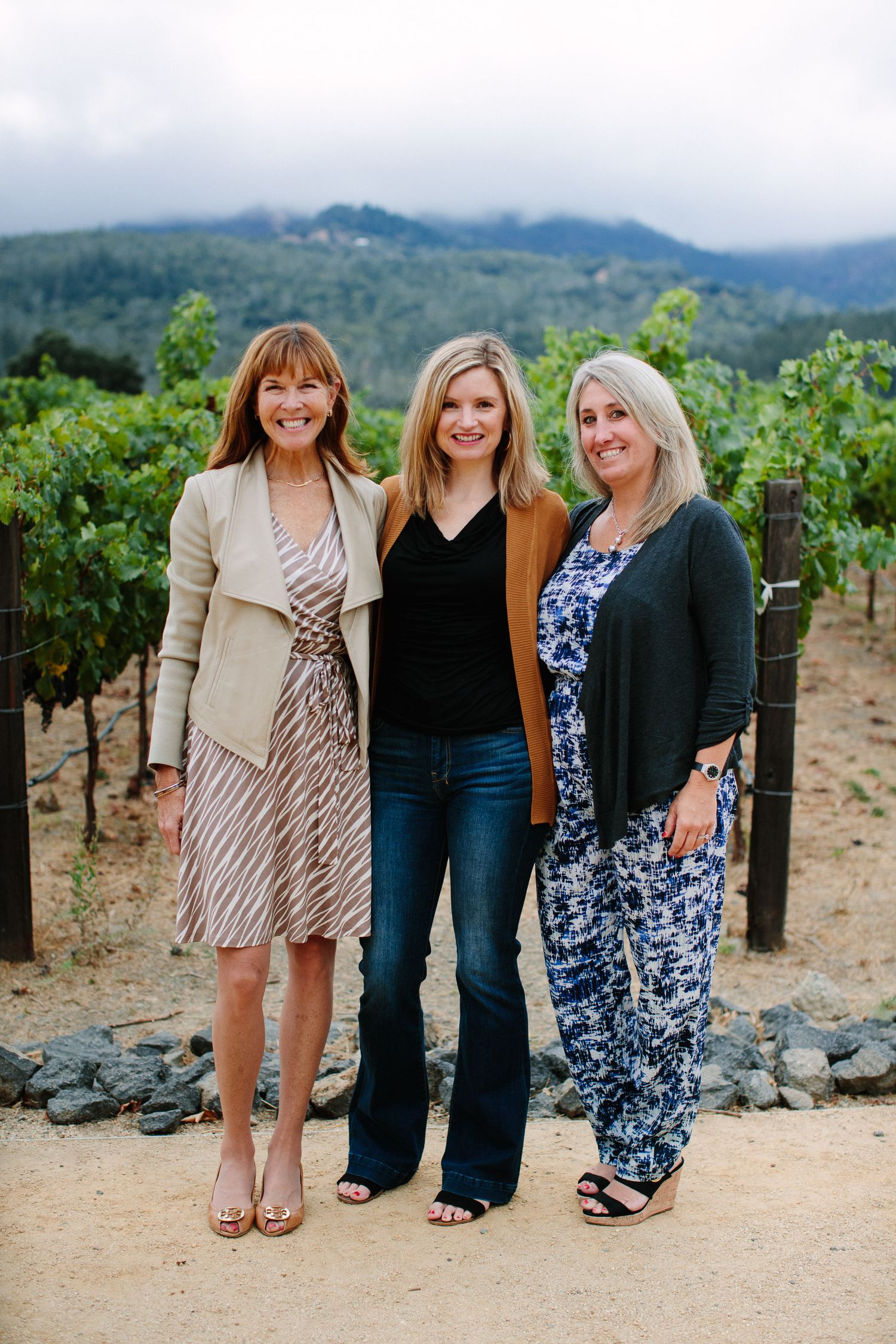 Three women stand together smiling at a vineyard with mountains visible in the background.