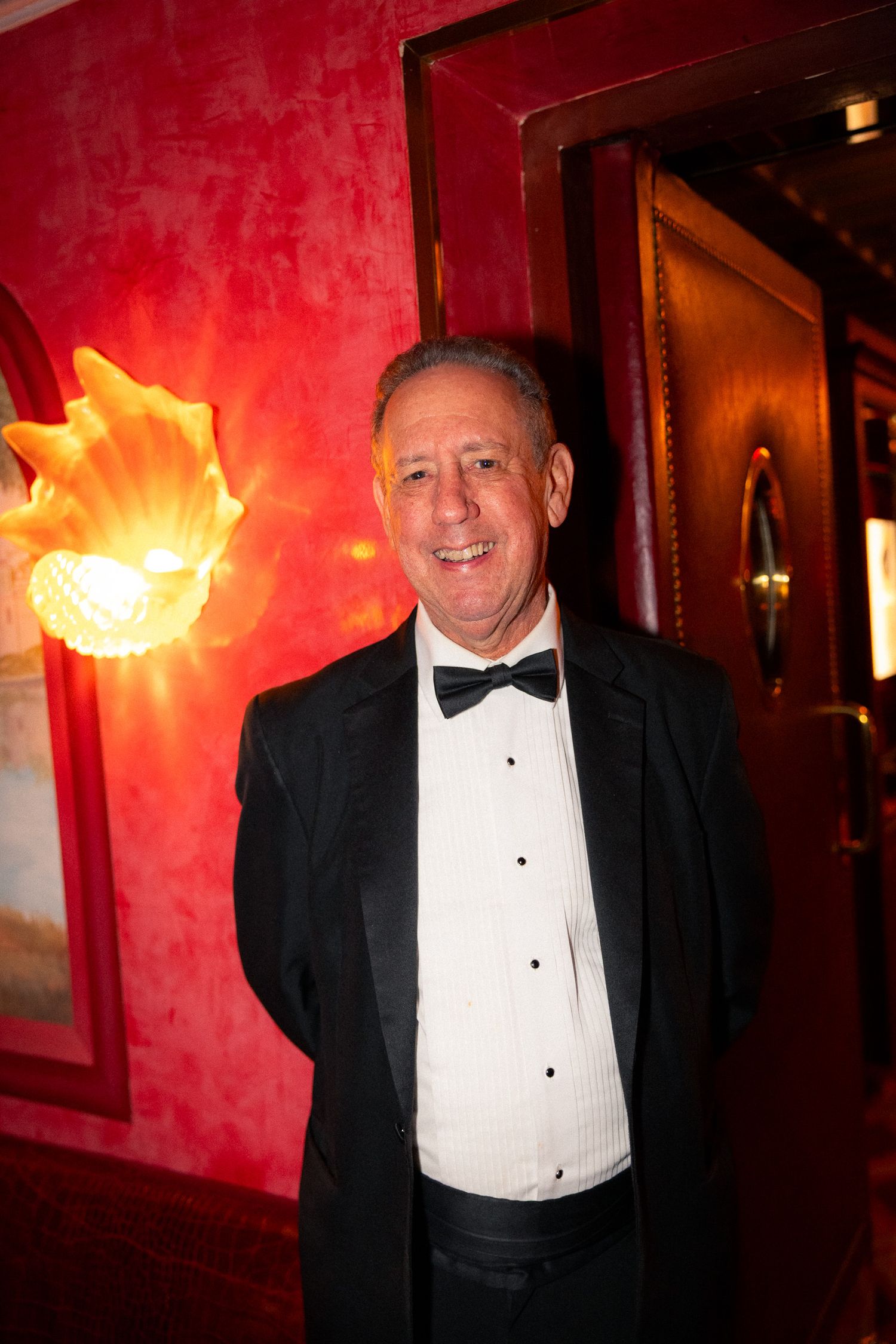 A person in formal black tuxedo and bow tie poses against a red wall with decorative lighting.