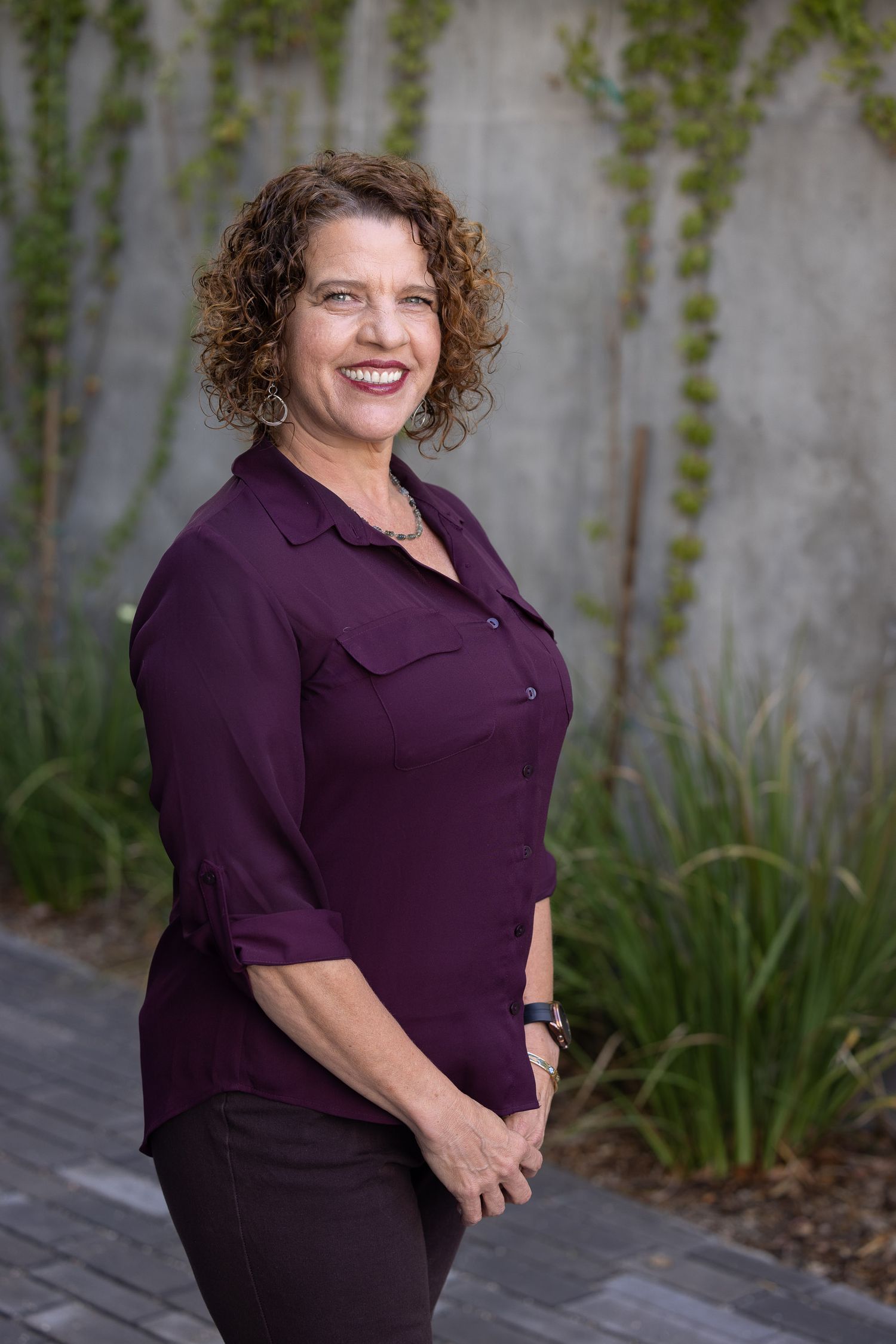 Professional portrait of person in purple button-up blouse standing against ivy covered wall.