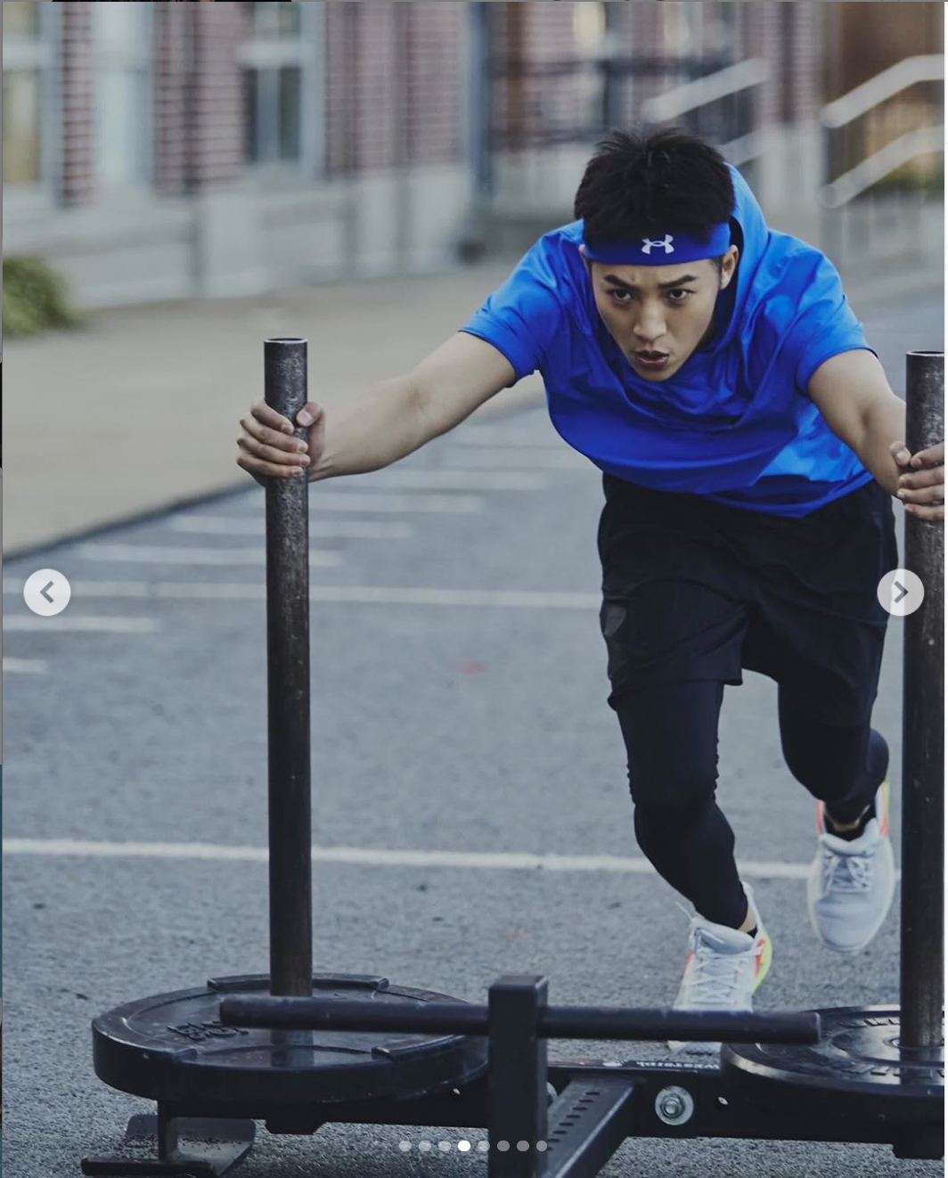 An athlete in a blue shirt and black pants pushes a weighted sled during an intense workout on pavement.