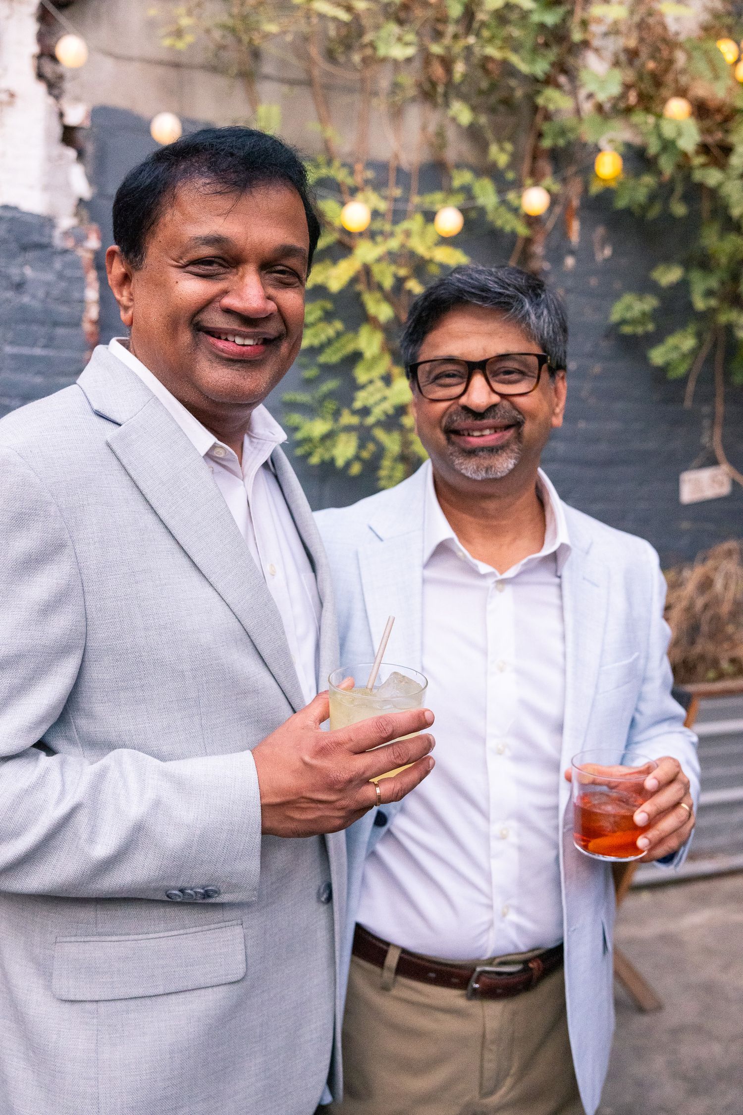 Two men in business attire enjoying drinks at an outdoor gathering with string lights.