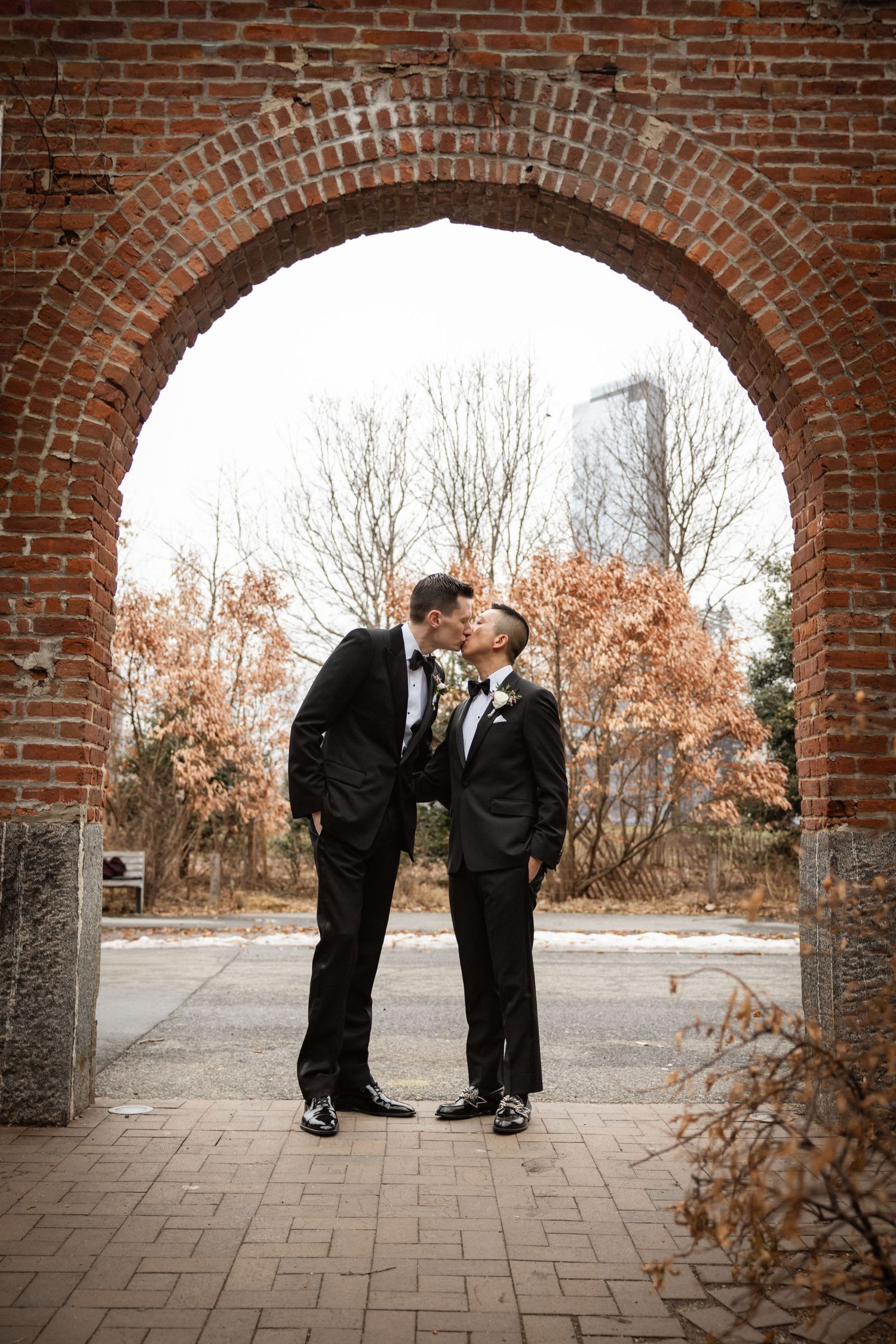 Two grooms in black tuxedos share a kiss under a red brick arch outdoors on their wedding day.