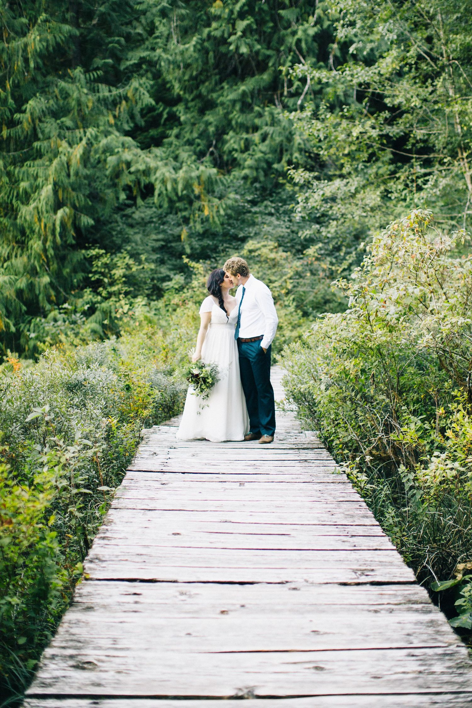 A couple stands on a rustic wooden bridge surrounded by lush green forest scenery during a wedding shoot.