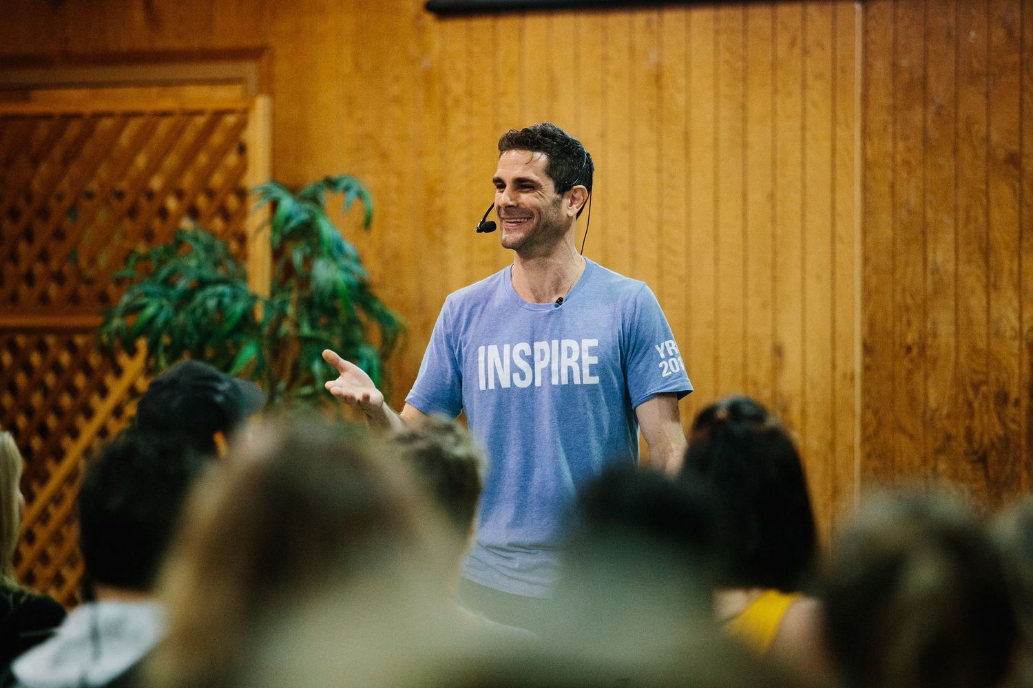 A person wearing a blue t-shirt speaks to a crowd at an outdoor gathering with wooden walls in the background.