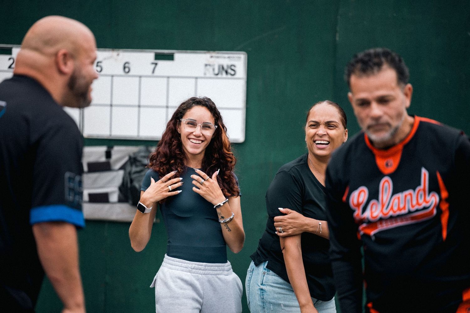 A lively group conversation between people standing near a scoreboard at a baseball diamond.