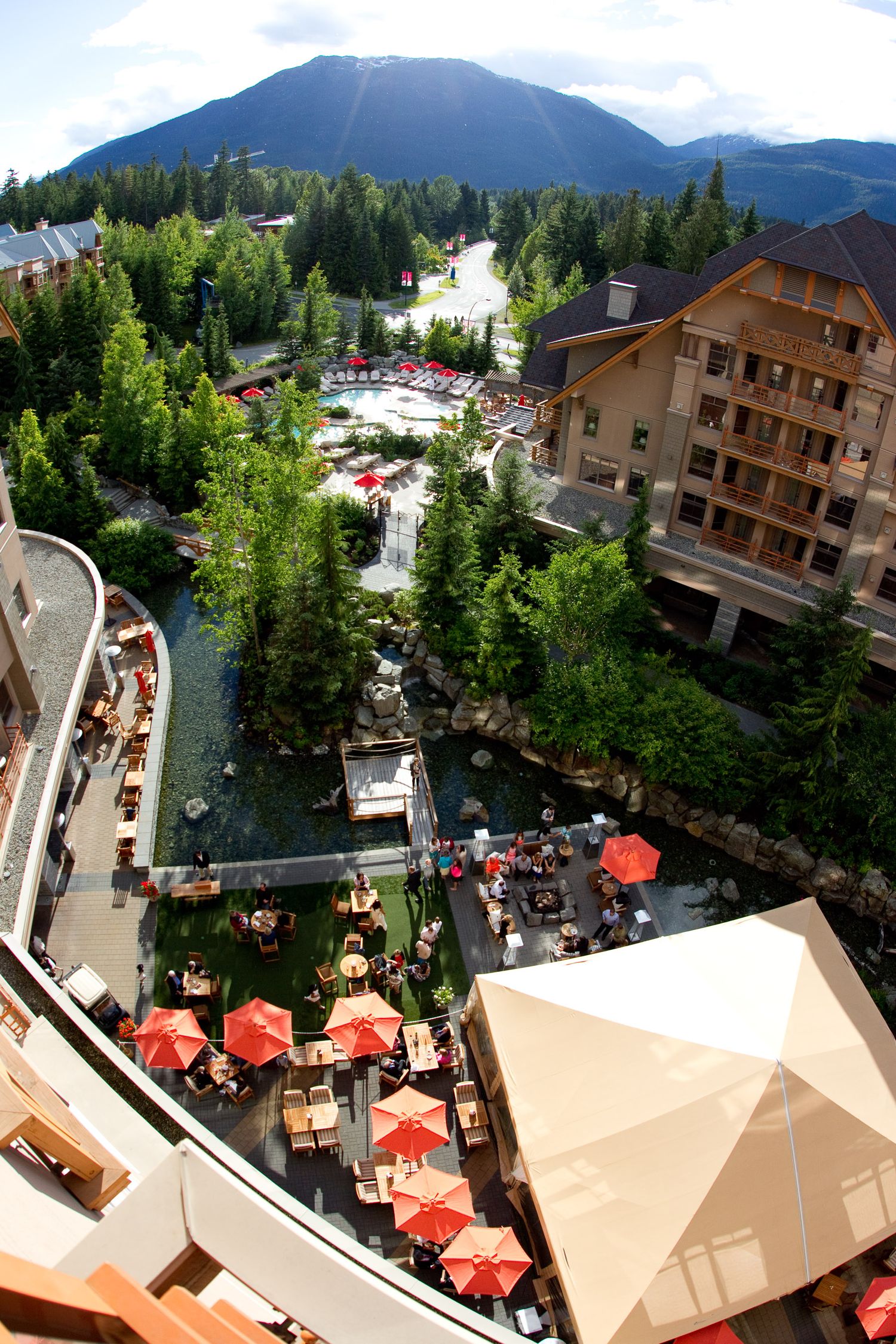 Aerial view of a mountain resort with red umbrellas and outdoor dining areas surrounded by evergreen trees.